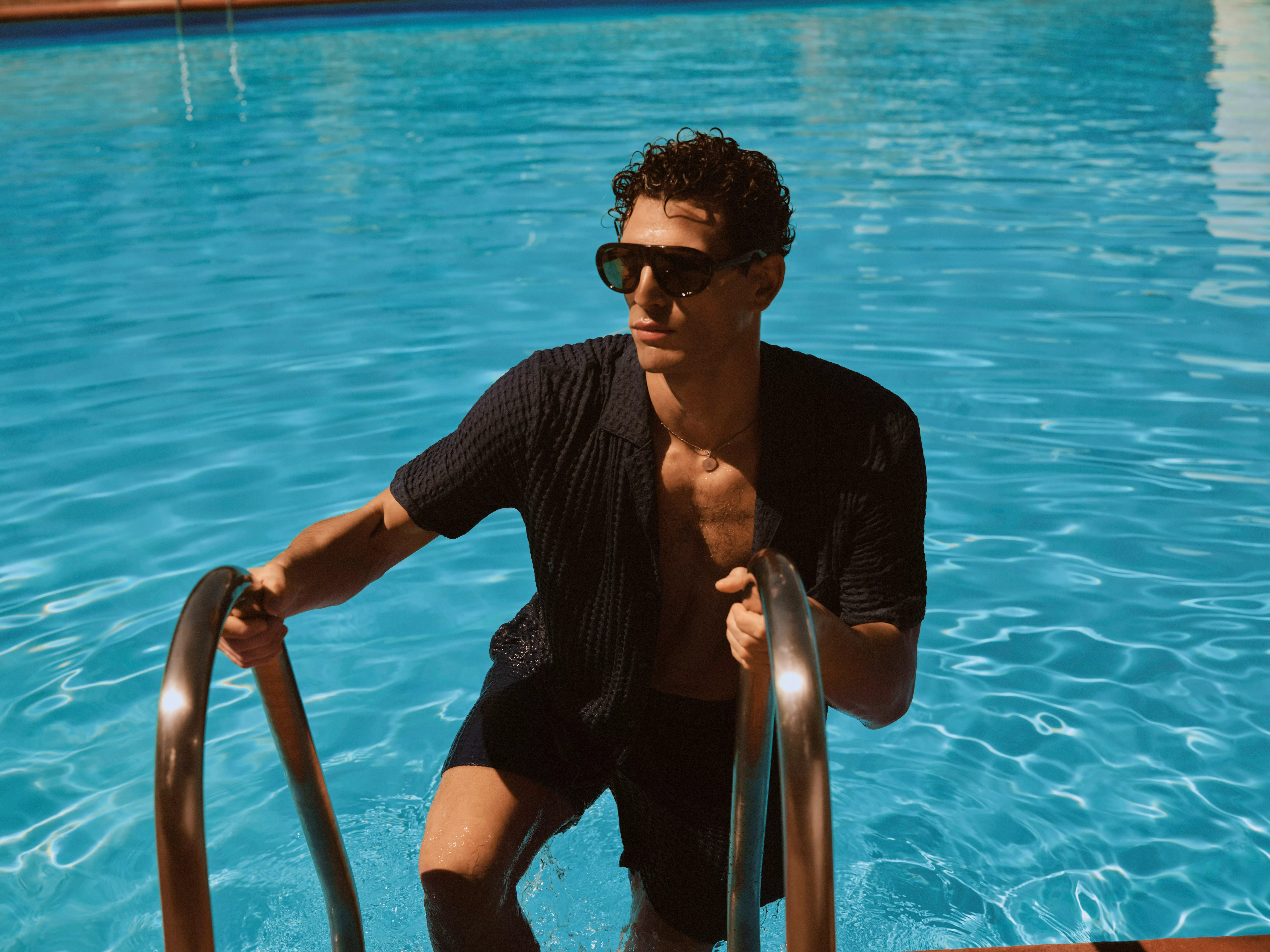 Man wearing sunglasses climbs pool ladder in bright blue swimming pool, summer holiday lifestyle portrait outdoors