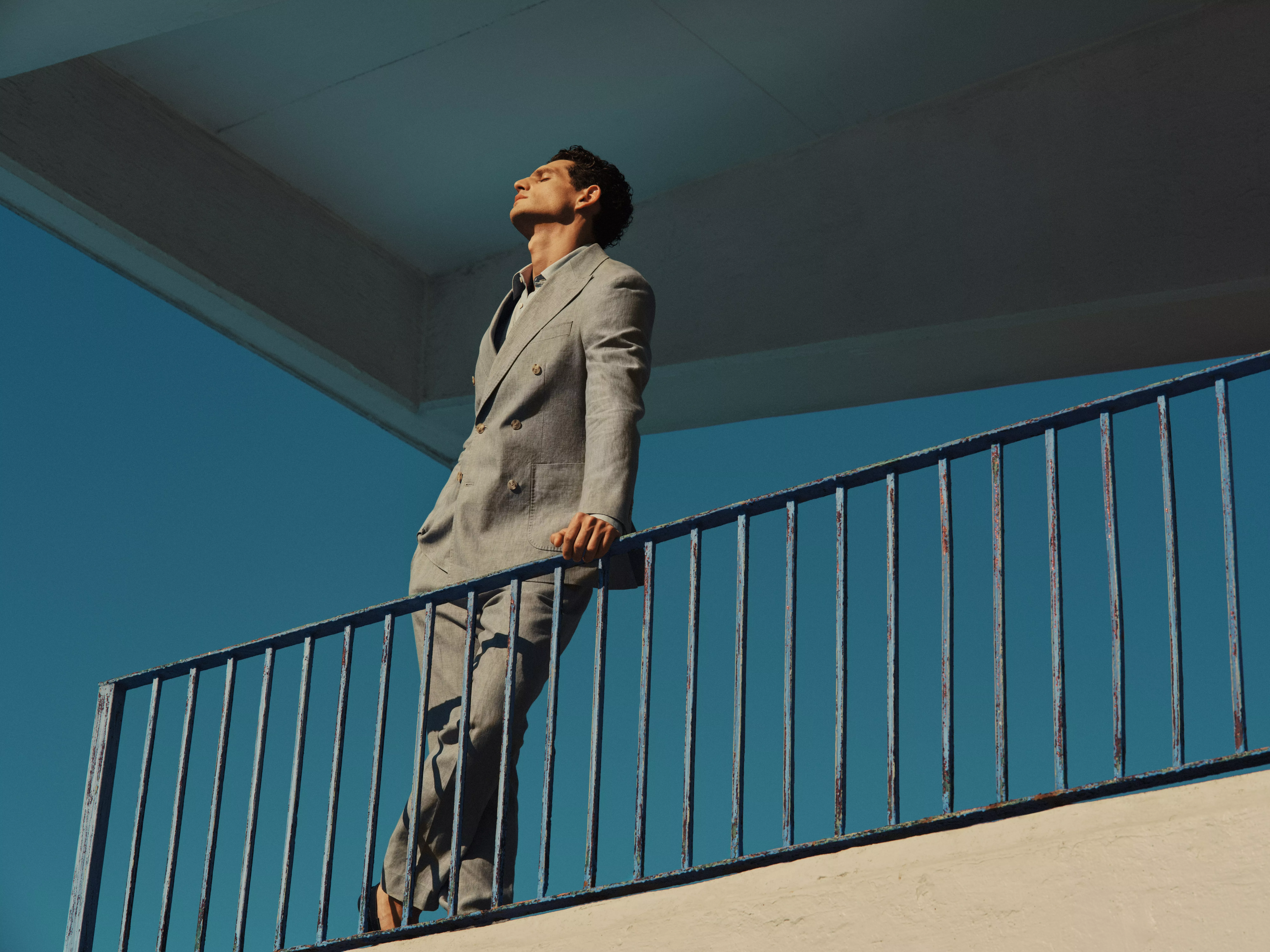 Man in light grey suit on balcony, leaning on blue railing under modern concrete roof, fashion portrait outdoors