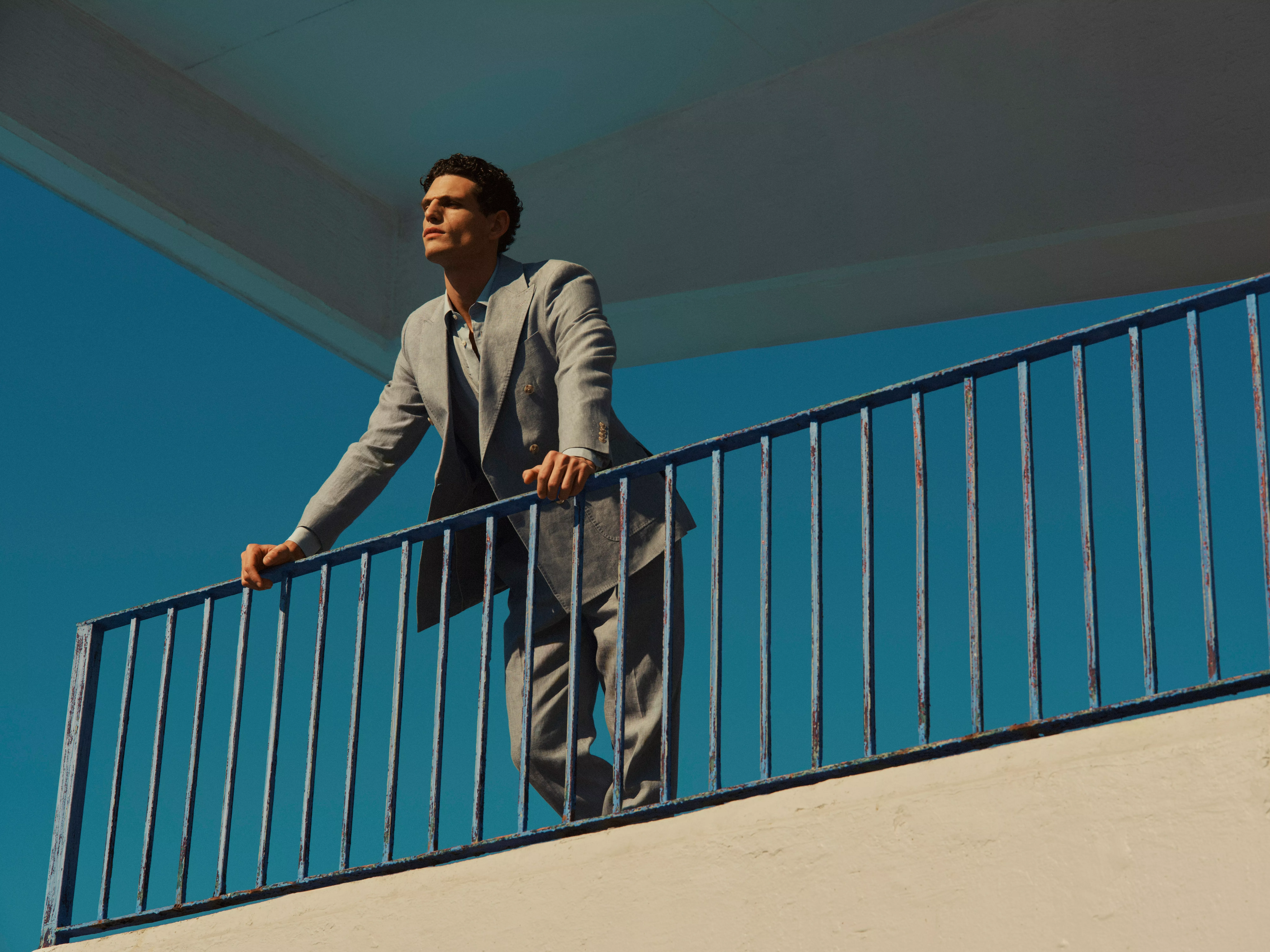 Stylish man in a light grey suit leaning on blue railings outdoors, modern fashion portrait against clear sky