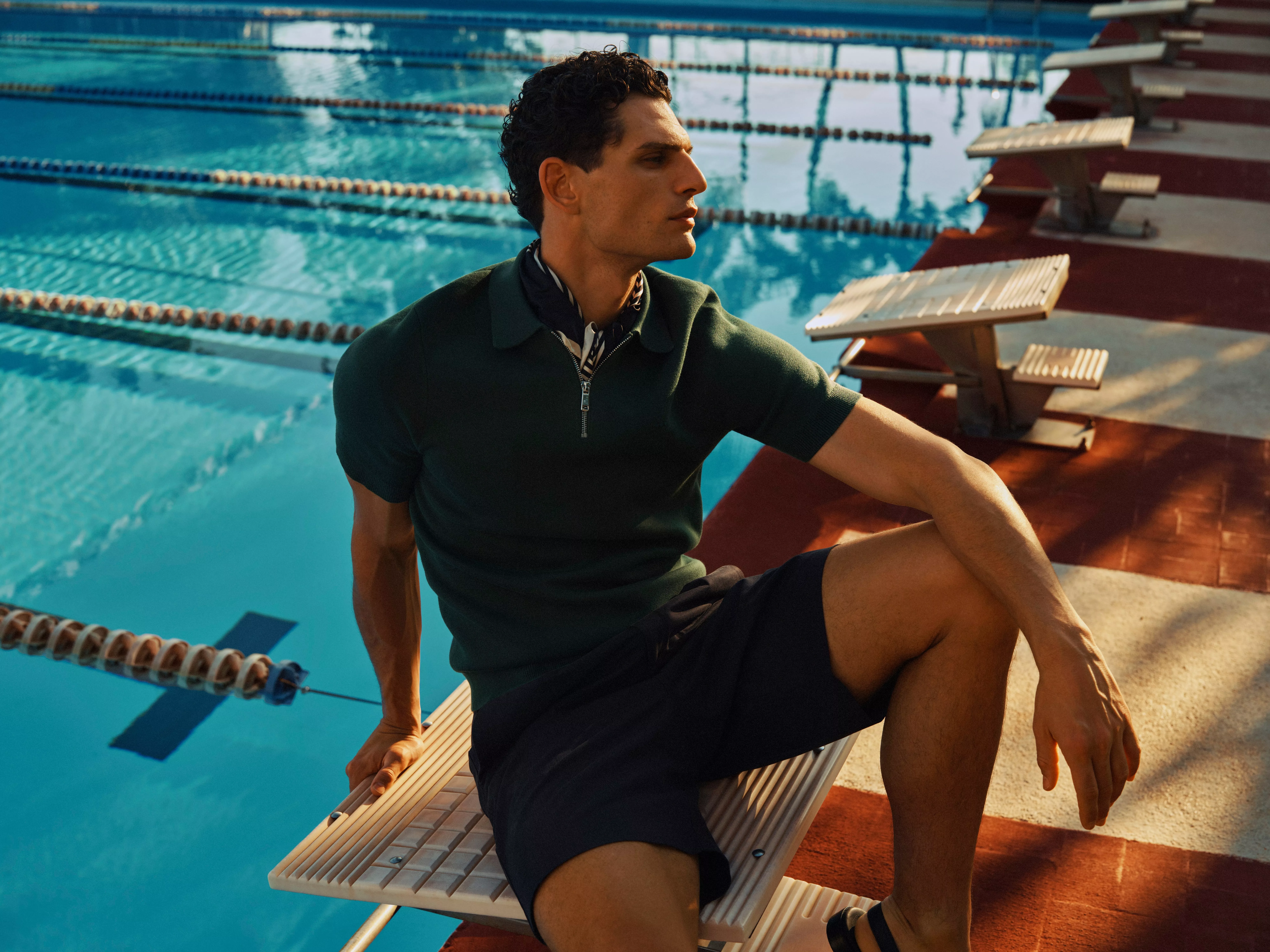 Man in smart casual polo and shorts sitting on a swimming pool starting block, outdoor sports lifestyle fashion shoot