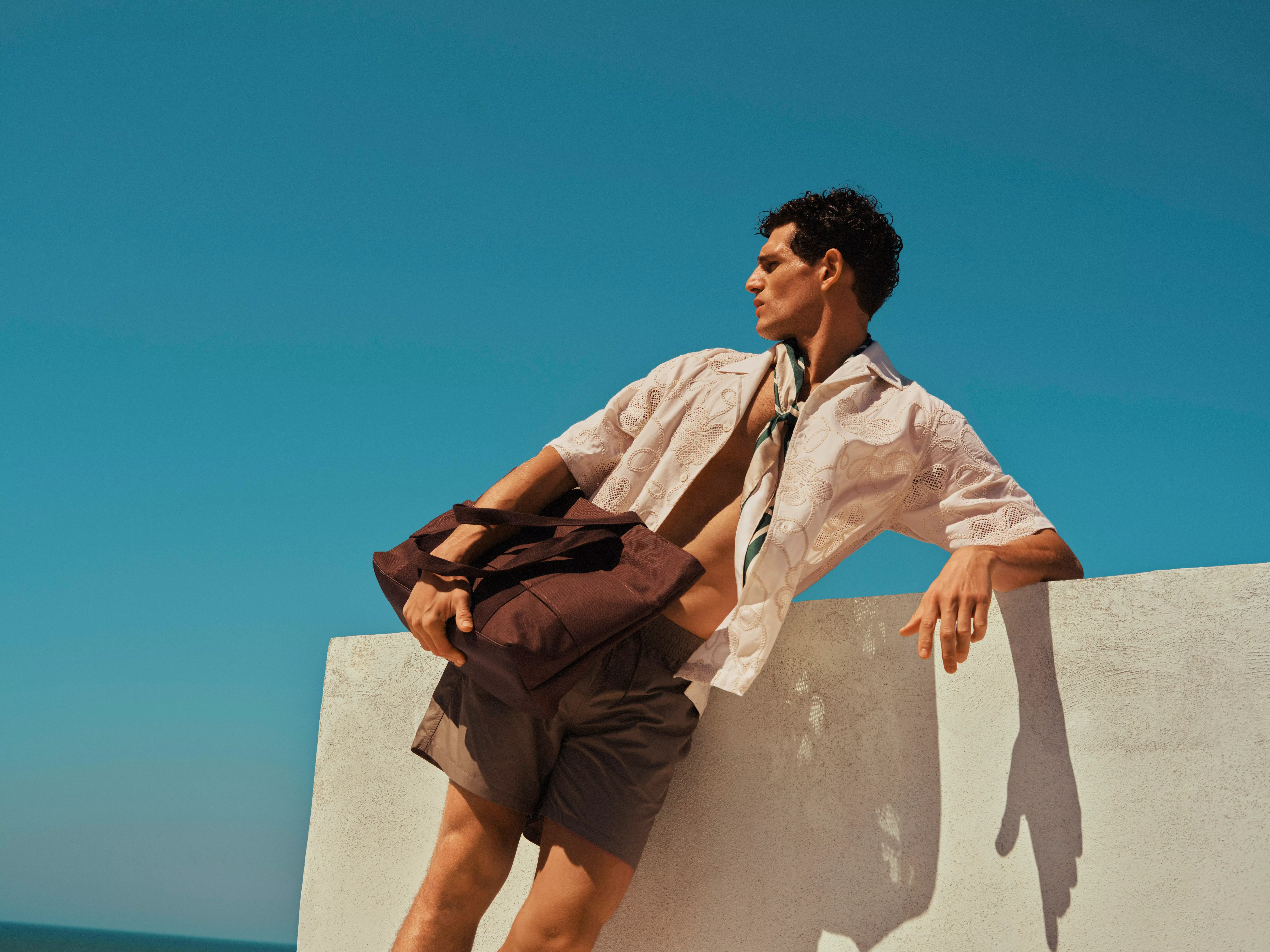 Stylish man in embroidered shirt and shorts holding a brown tote bag, leaning on a sunlit wall under blue sky