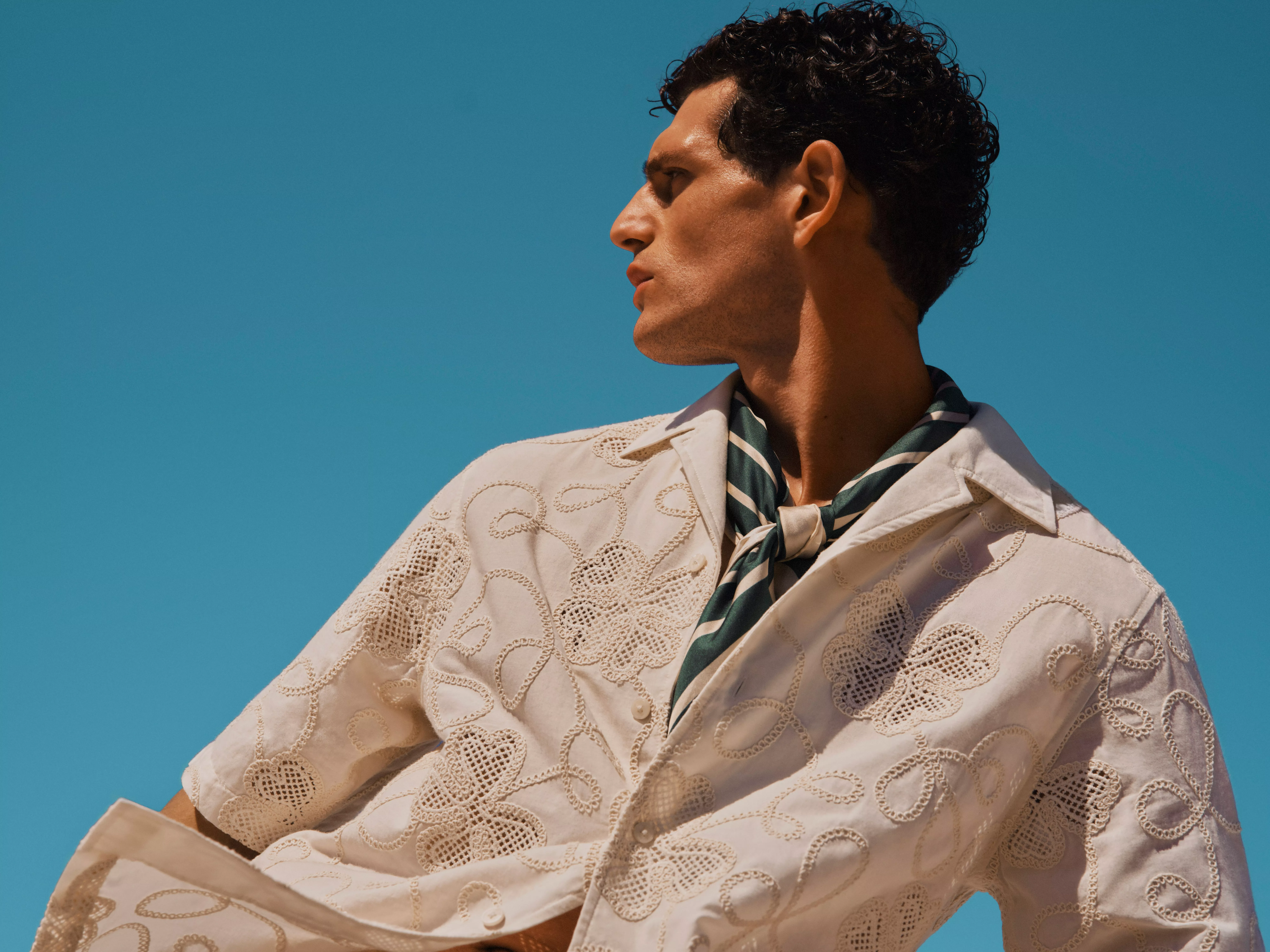 Man in embroidered cream short-sleeve shirt and striped neck scarf, looking sideways under clear blue sky