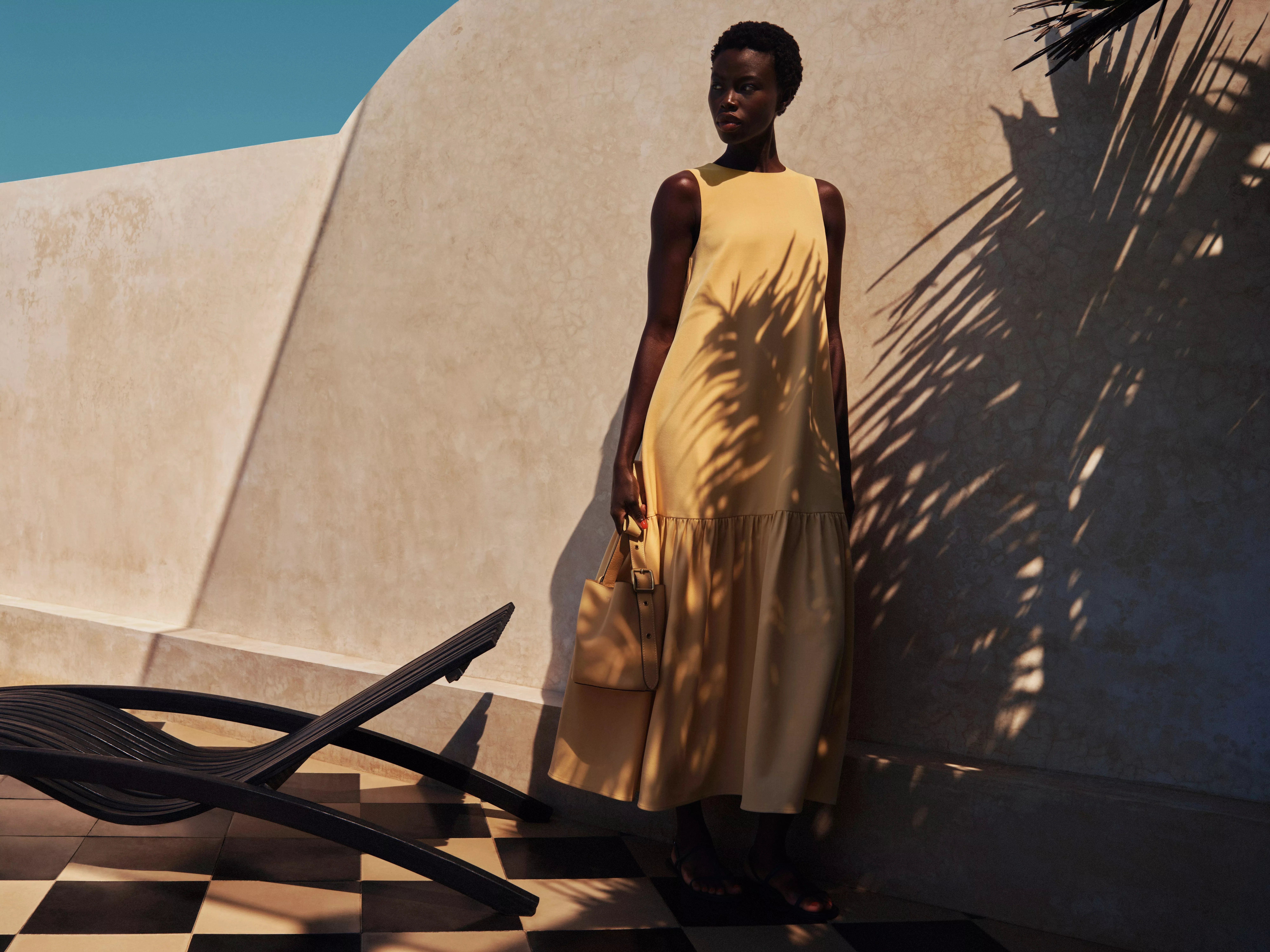 Fashion model in a mustard maxi dress holding a tote bag, posed by a sunlit wall with palm shadows outdoors
