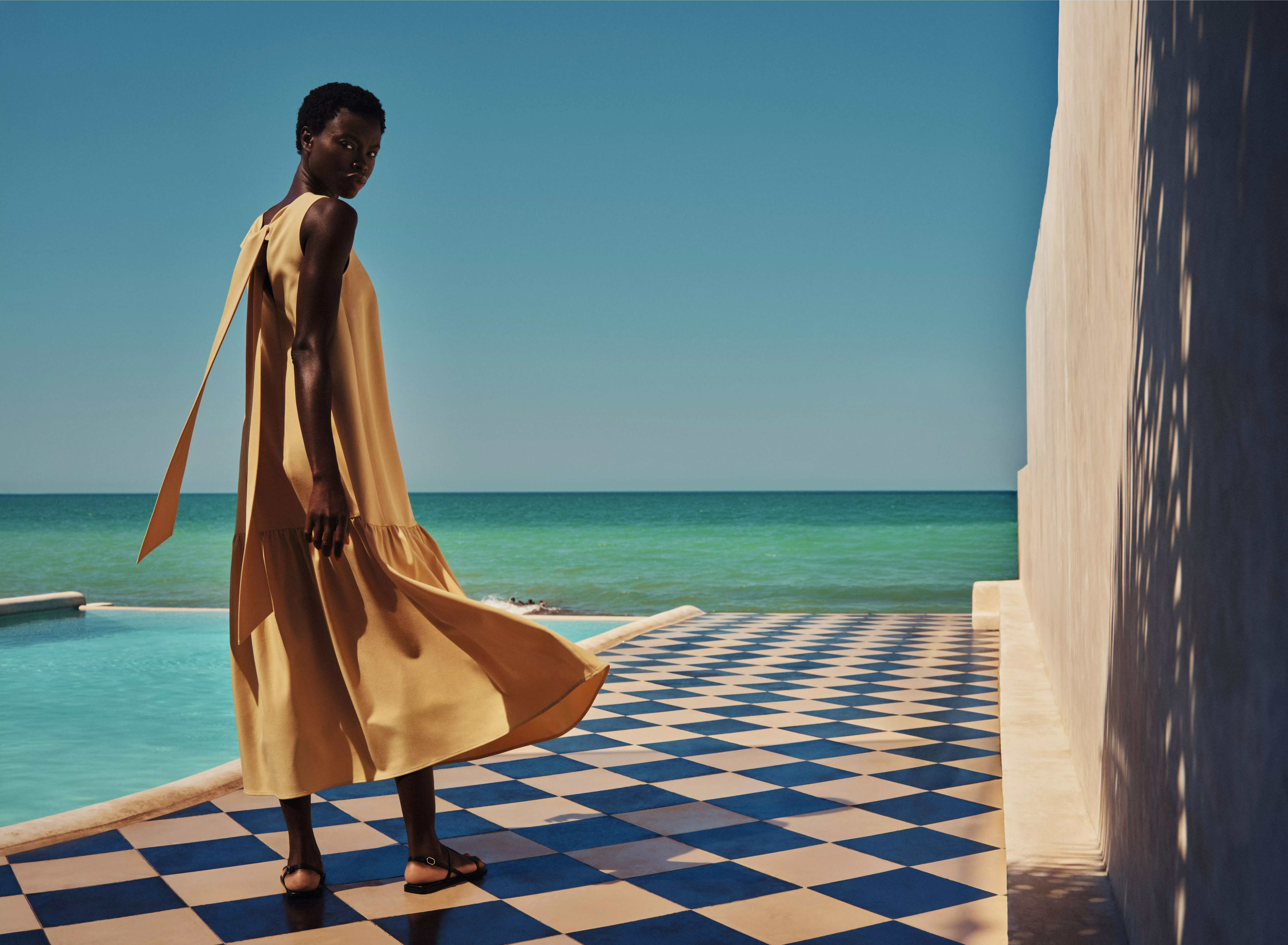 Fashion model in flowing mustard maxi dress by turquoise sea, on blue-and-white tiled terrace in sunlight