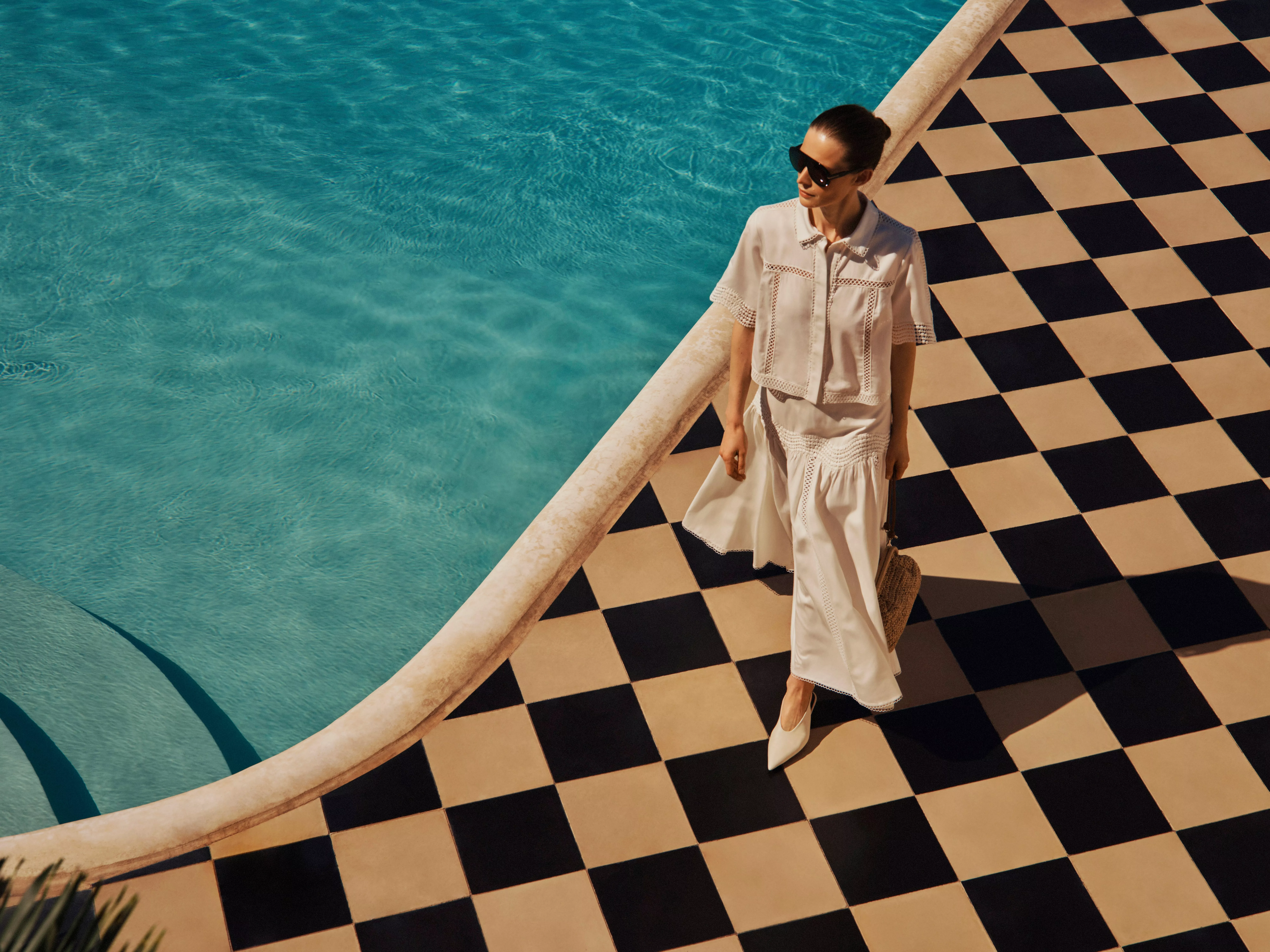 Woman in white summer top and skirt walking by pool on black and cream chequered tiles, luxury resort fashion scene