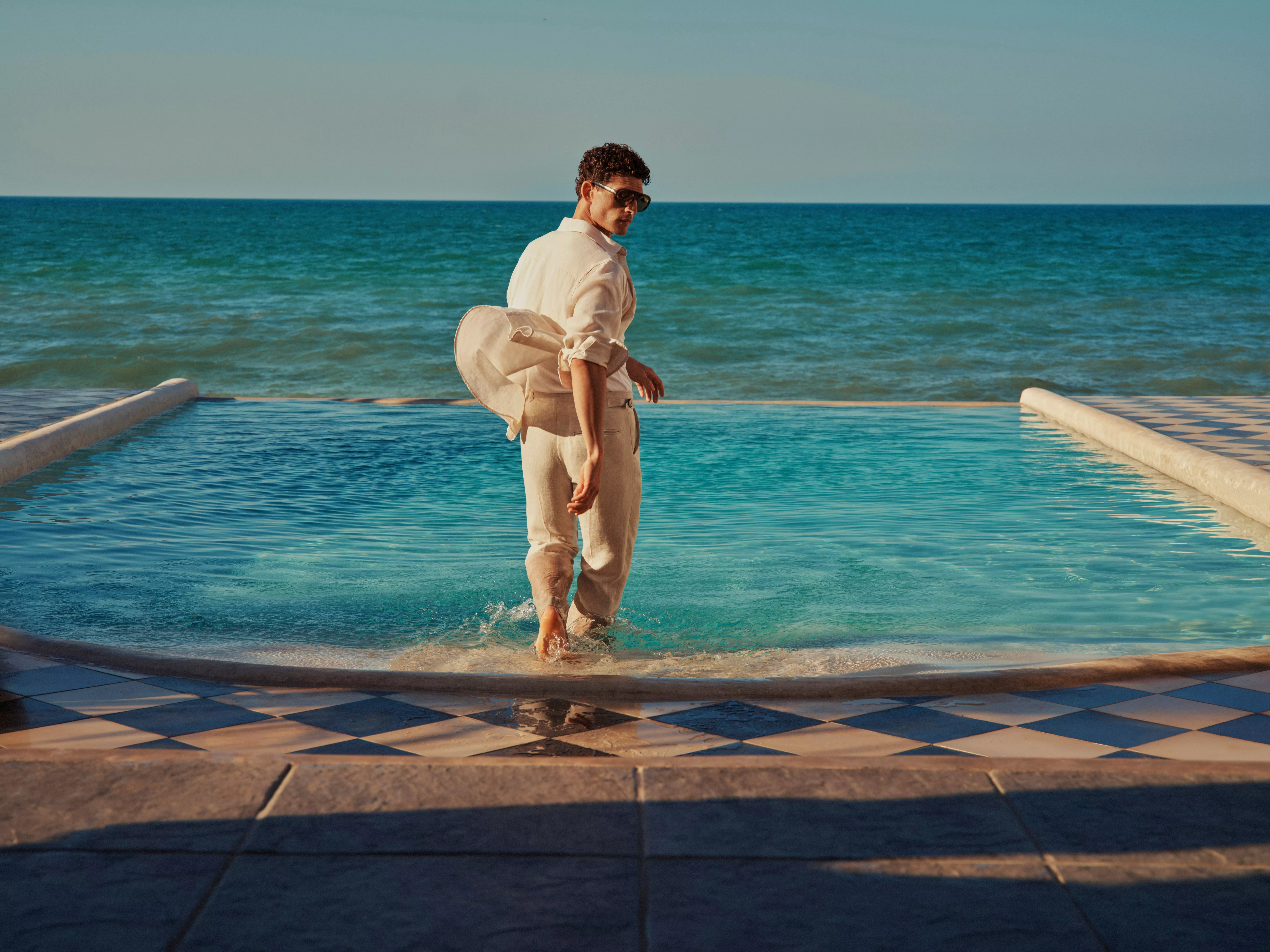 Man in linen shirt and trousers walking through an infinity pool by the sea, wearing sunglasses at sunset.