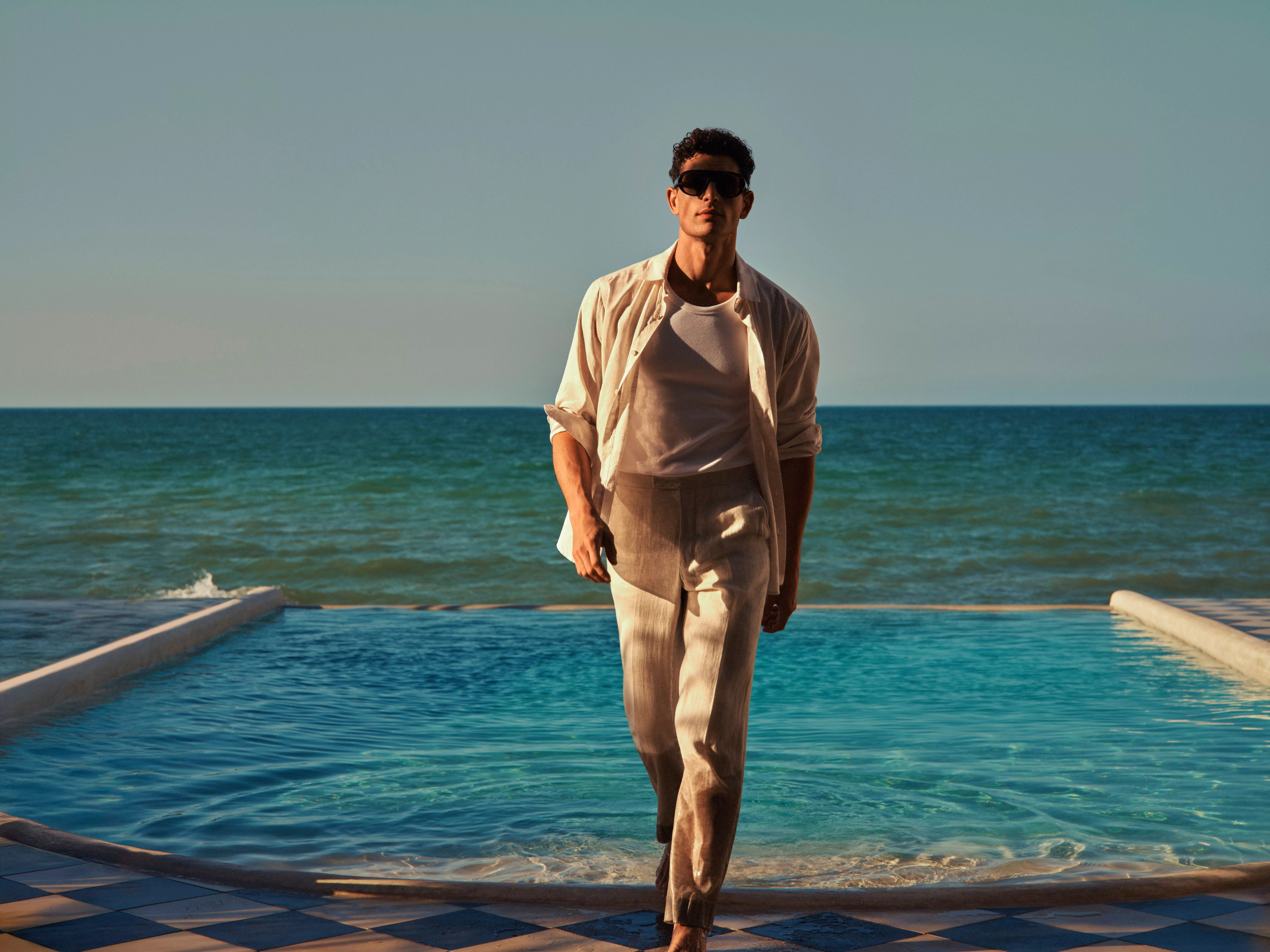 Stylish man in linen shirt and trousers walking by pool, with sea backdrop, wearing sunglasses at sunset