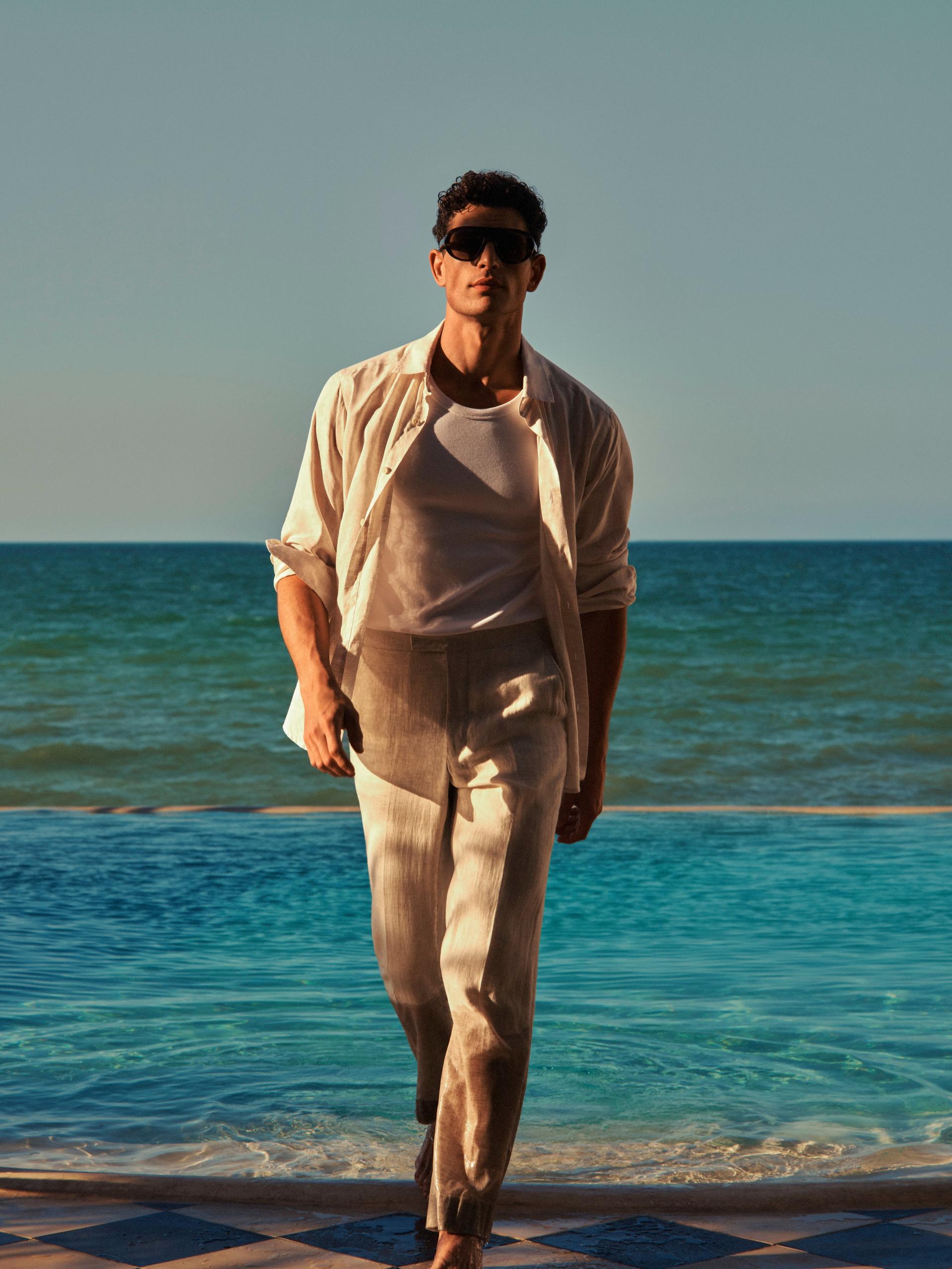Stylish man in linen shirt and trousers walking by pool, with sea backdrop, wearing sunglasses at sunset