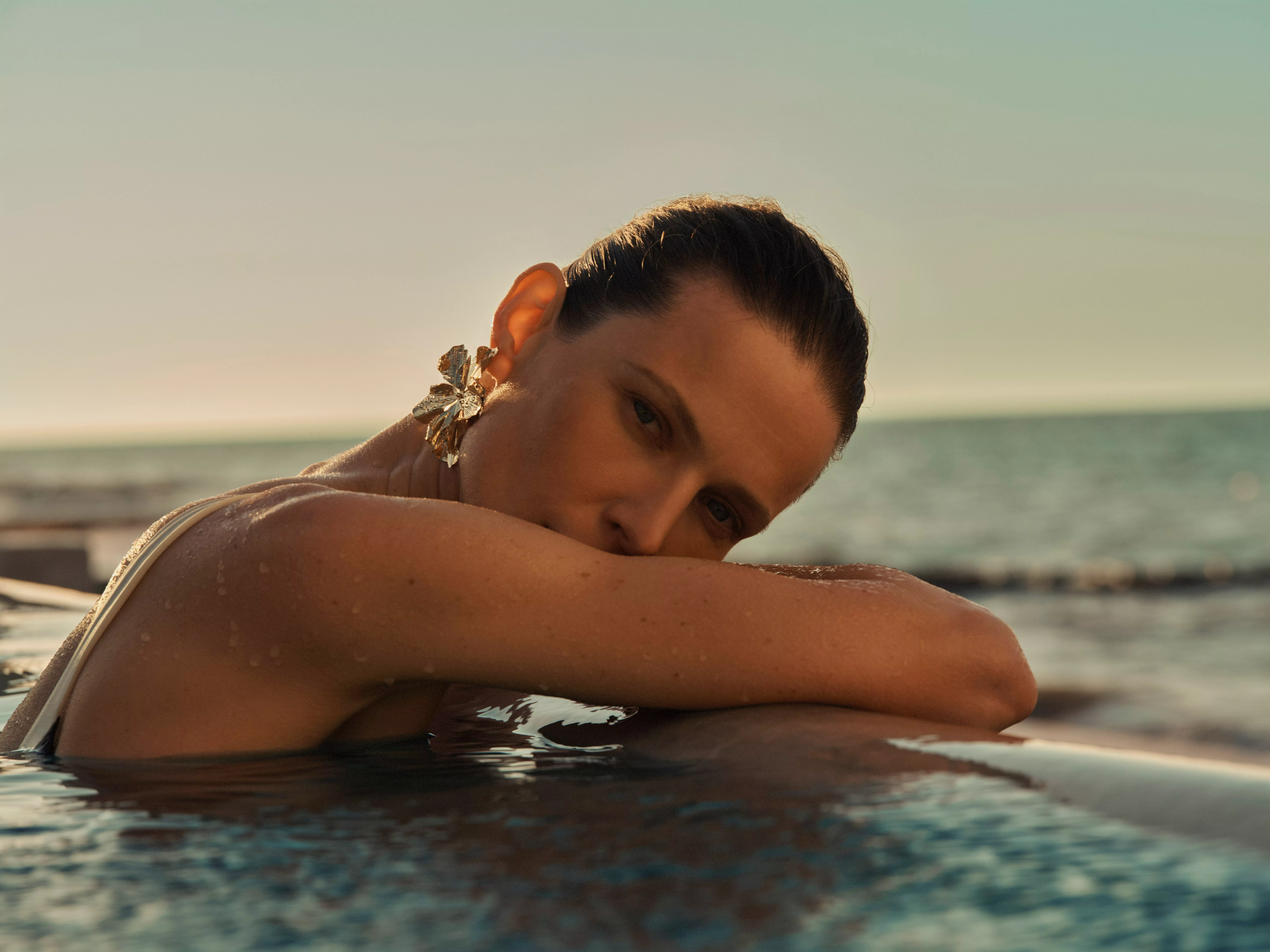 Woman relaxing in an infinity pool by the sea at sunset, wearing statement earrings and looking at camera