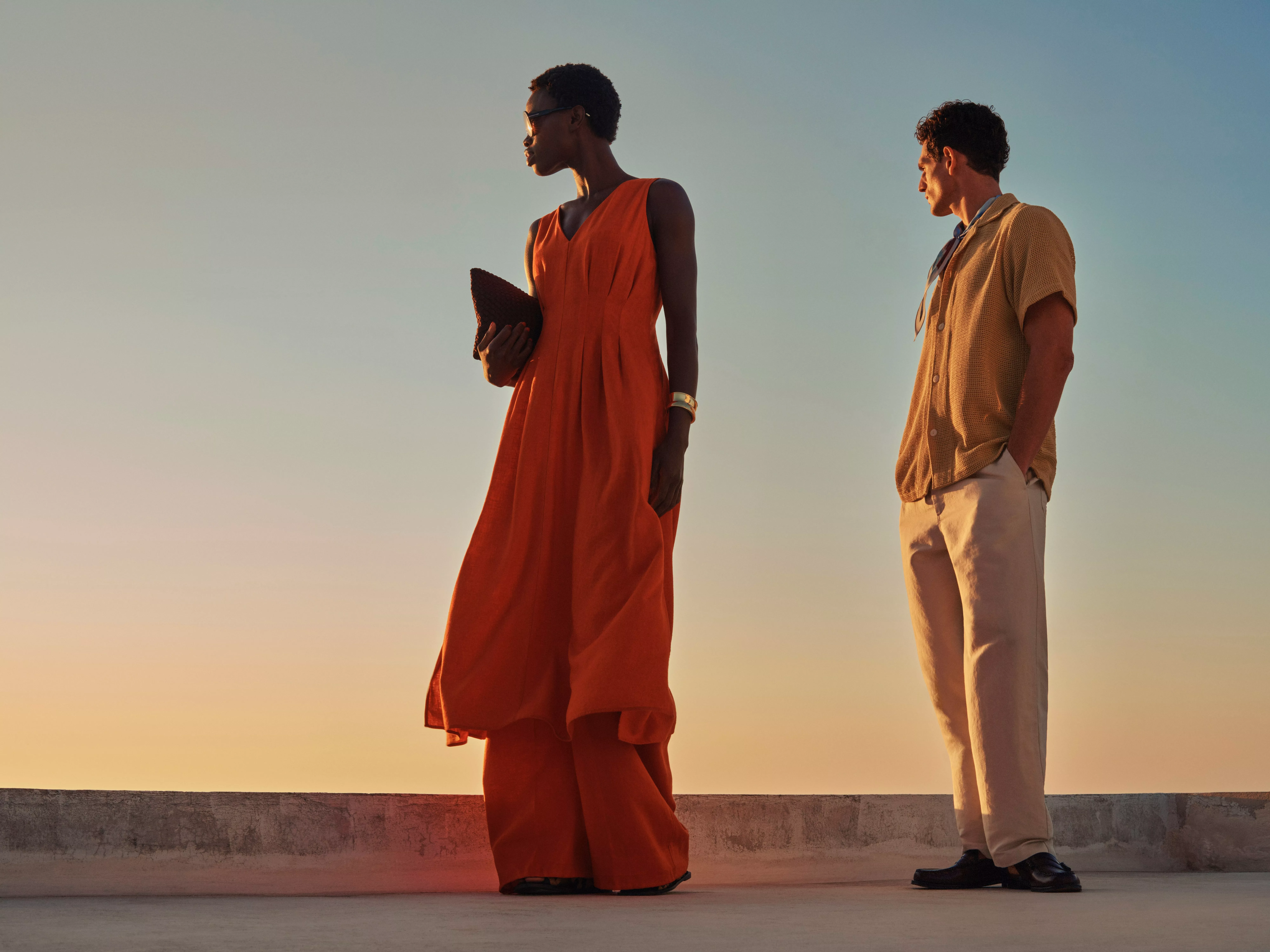 Two fashion models in summer outfits at sunset, woman in orange dress with clutch, man in beige shirt and trousers