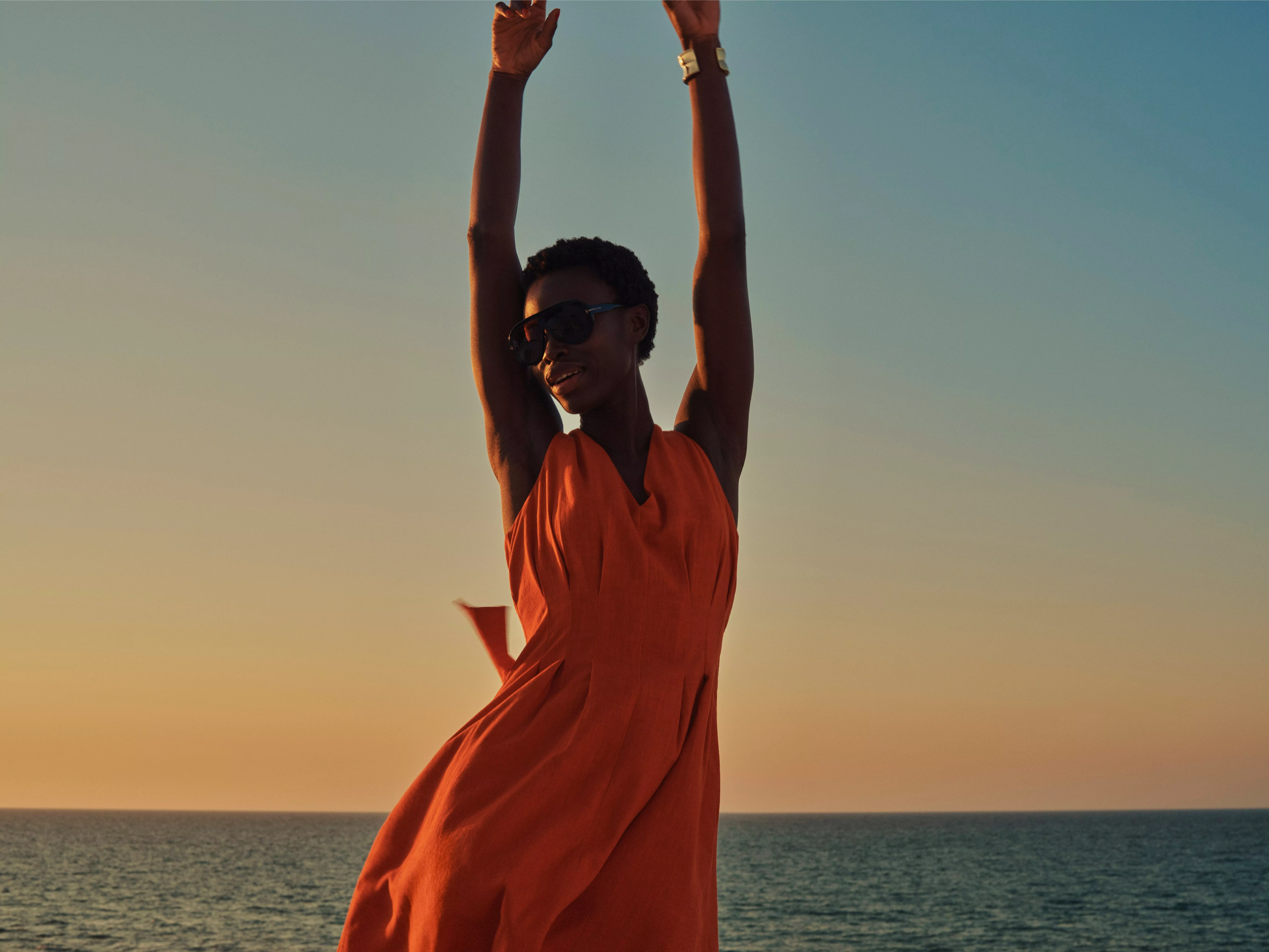 Woman in flowing orange dress and sunglasses raising arms at sunset by the sea, summer holiday lifestyle photo