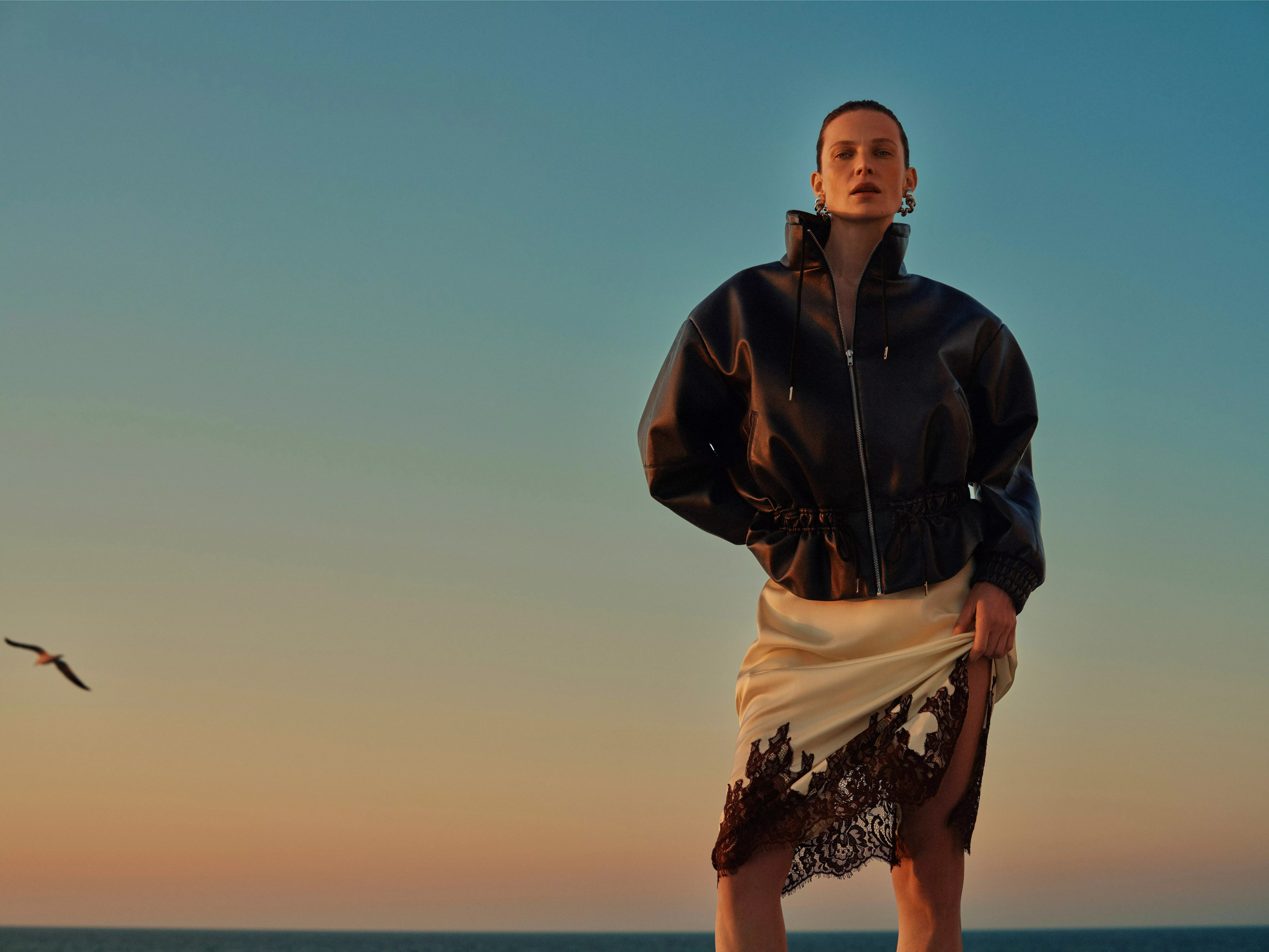 Fashion model in patterned camisole and denim Bermuda shorts, holding printed scarf against clear blue sky