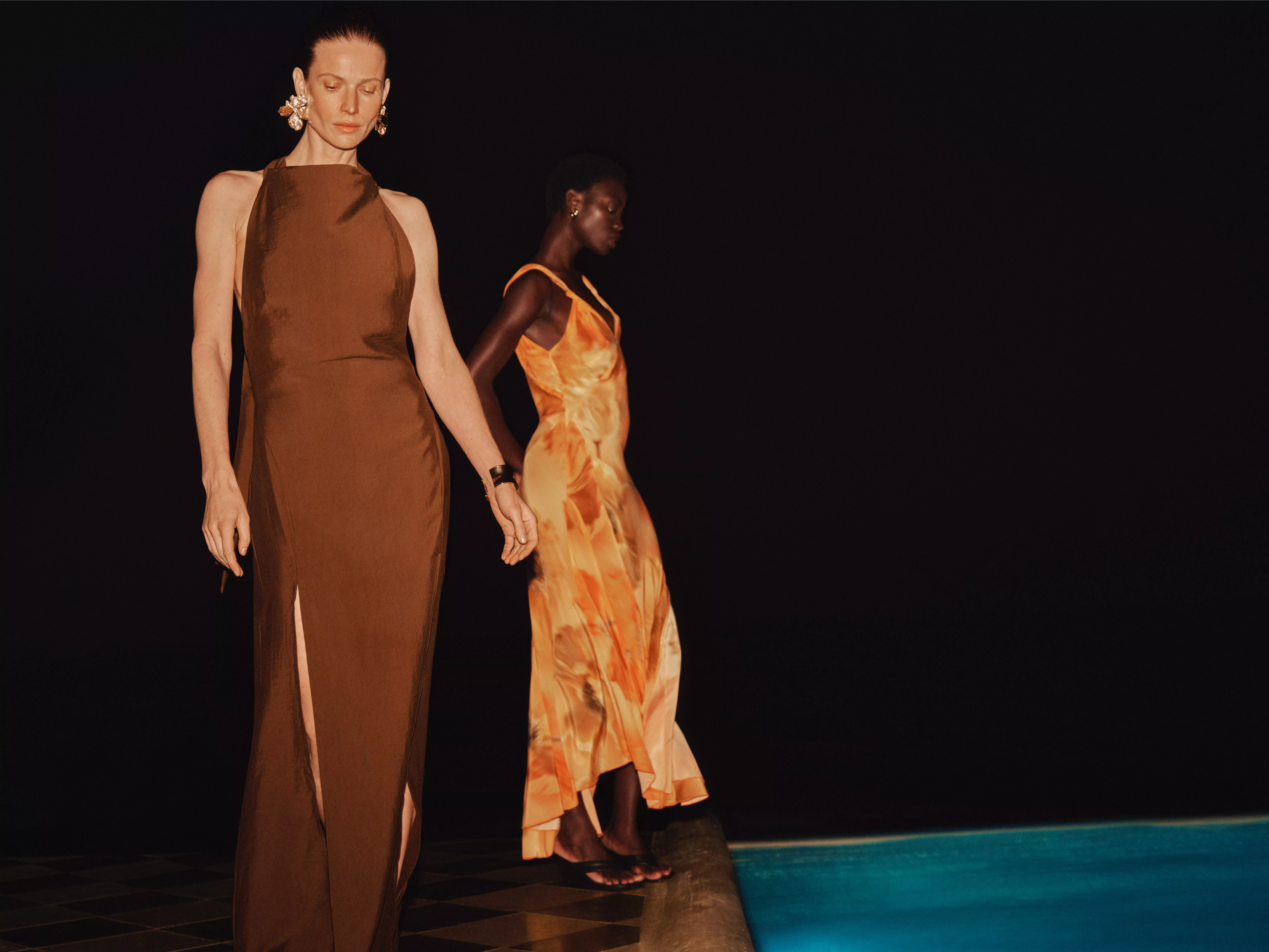 Two fashion models in elegant maxi dresses beside a pool, showcasing modern eveningwear style in low light