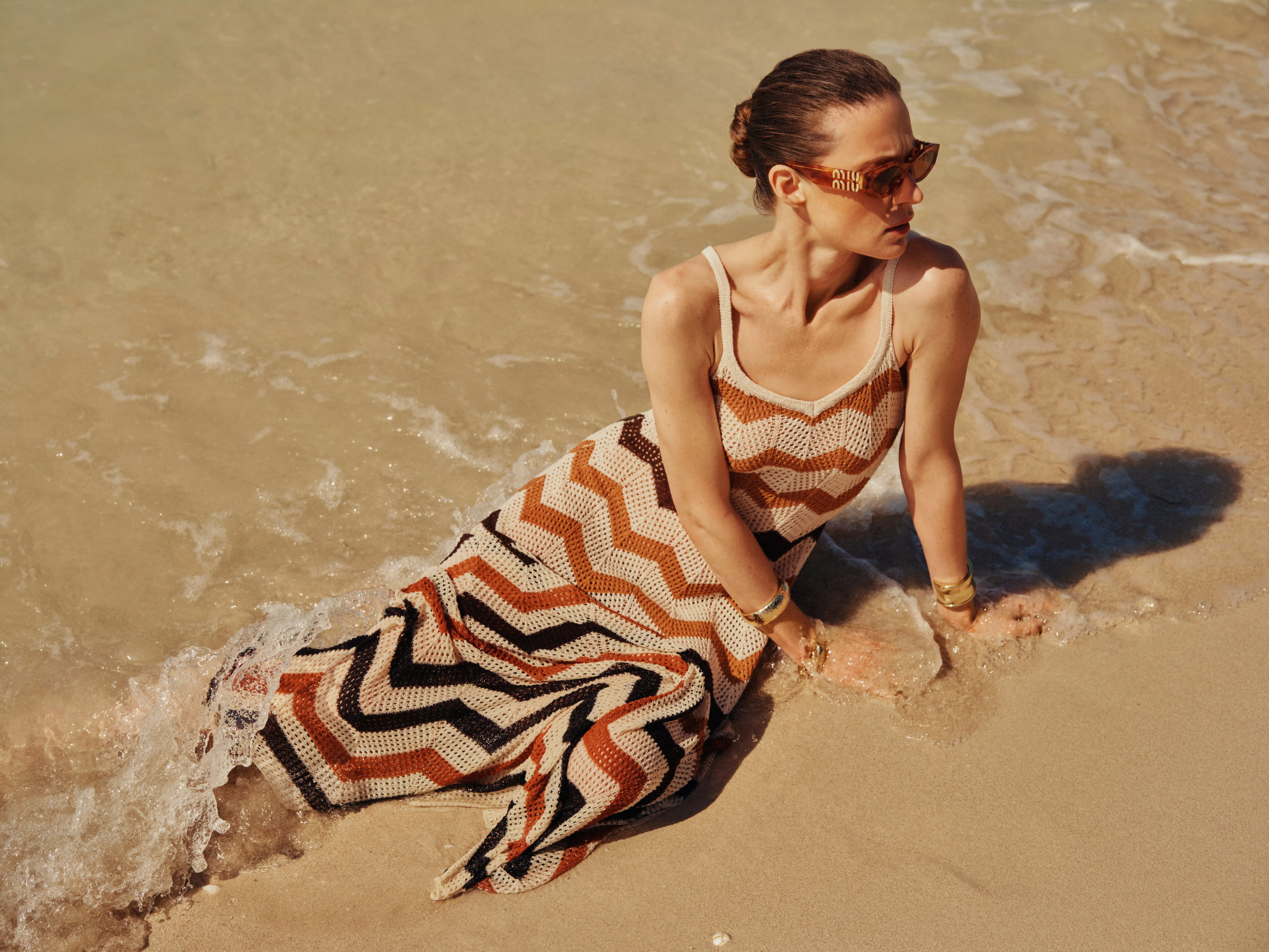 Woman in sunglasses wearing a zigzag knit maxi dress sitting in shallow sea water on a sandy beach.