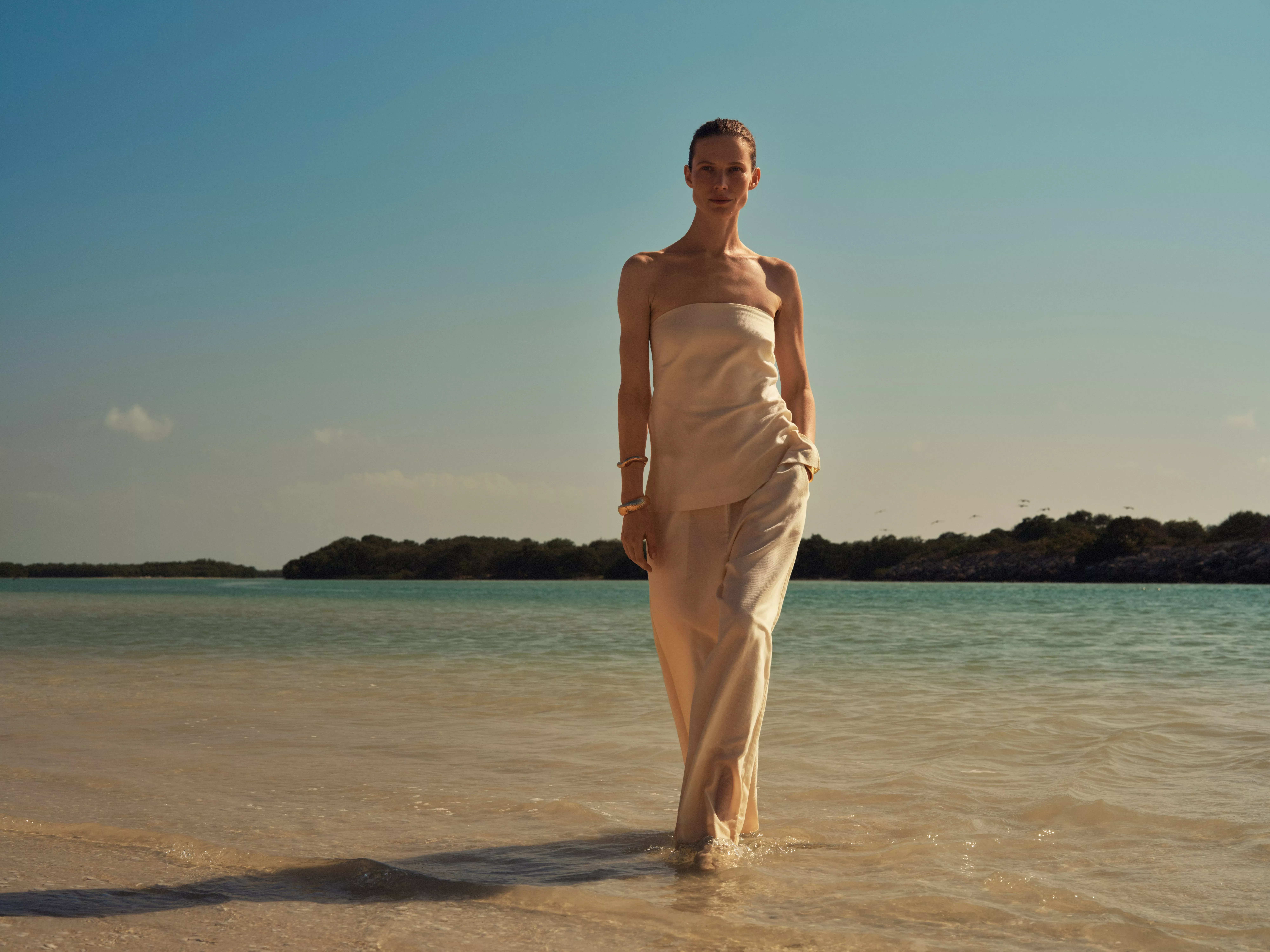 Woman in cream strapless top and satin trousers walking through shallow sea at a sunny tropical beach.