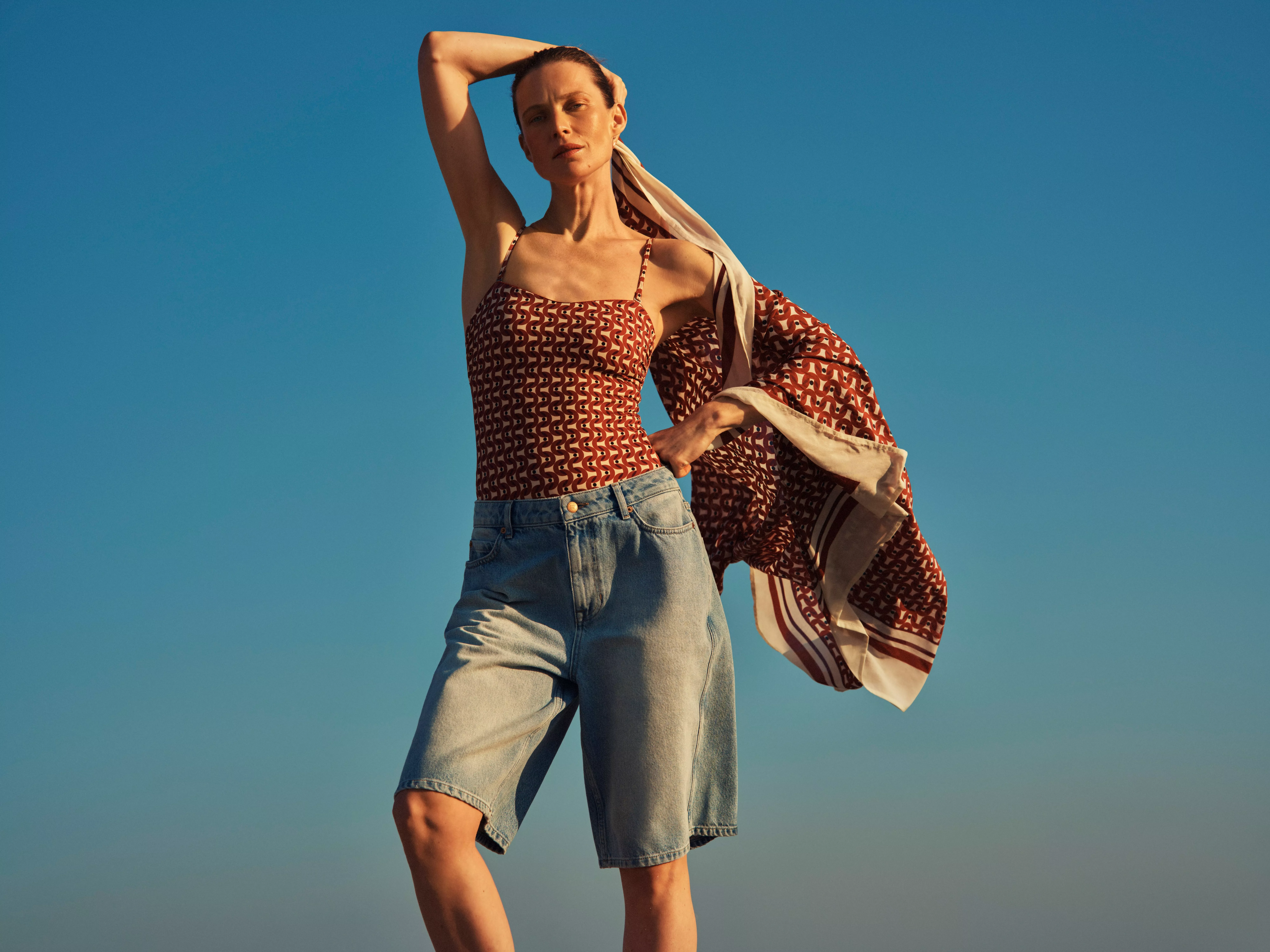 Fashion model in patterned camisole and denim Bermuda shorts, holding printed scarf against clear blue sky