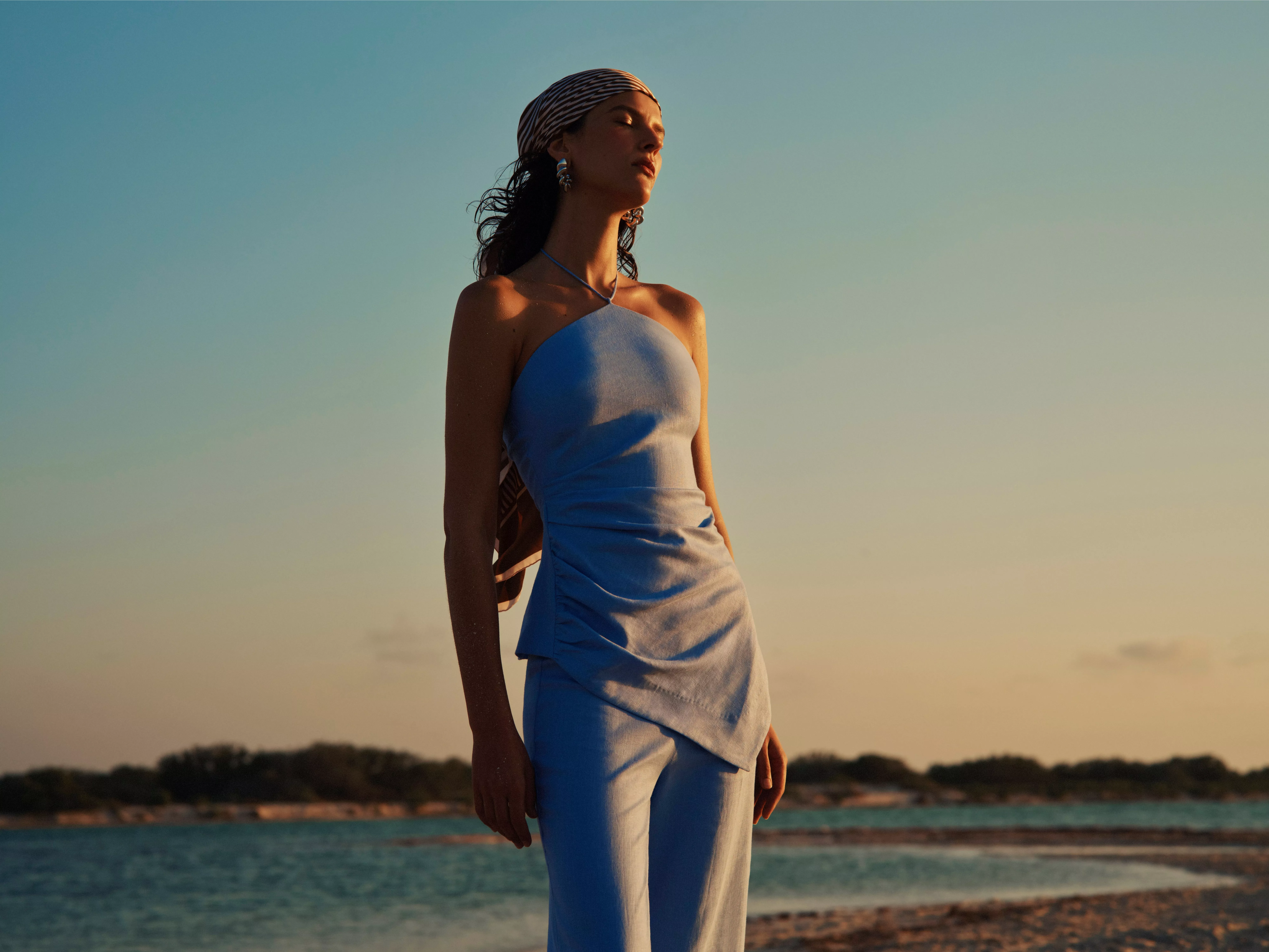 Woman in light blue outfit and headscarf posing by the sea at sunset, stylish summer fashion portrait outdoors