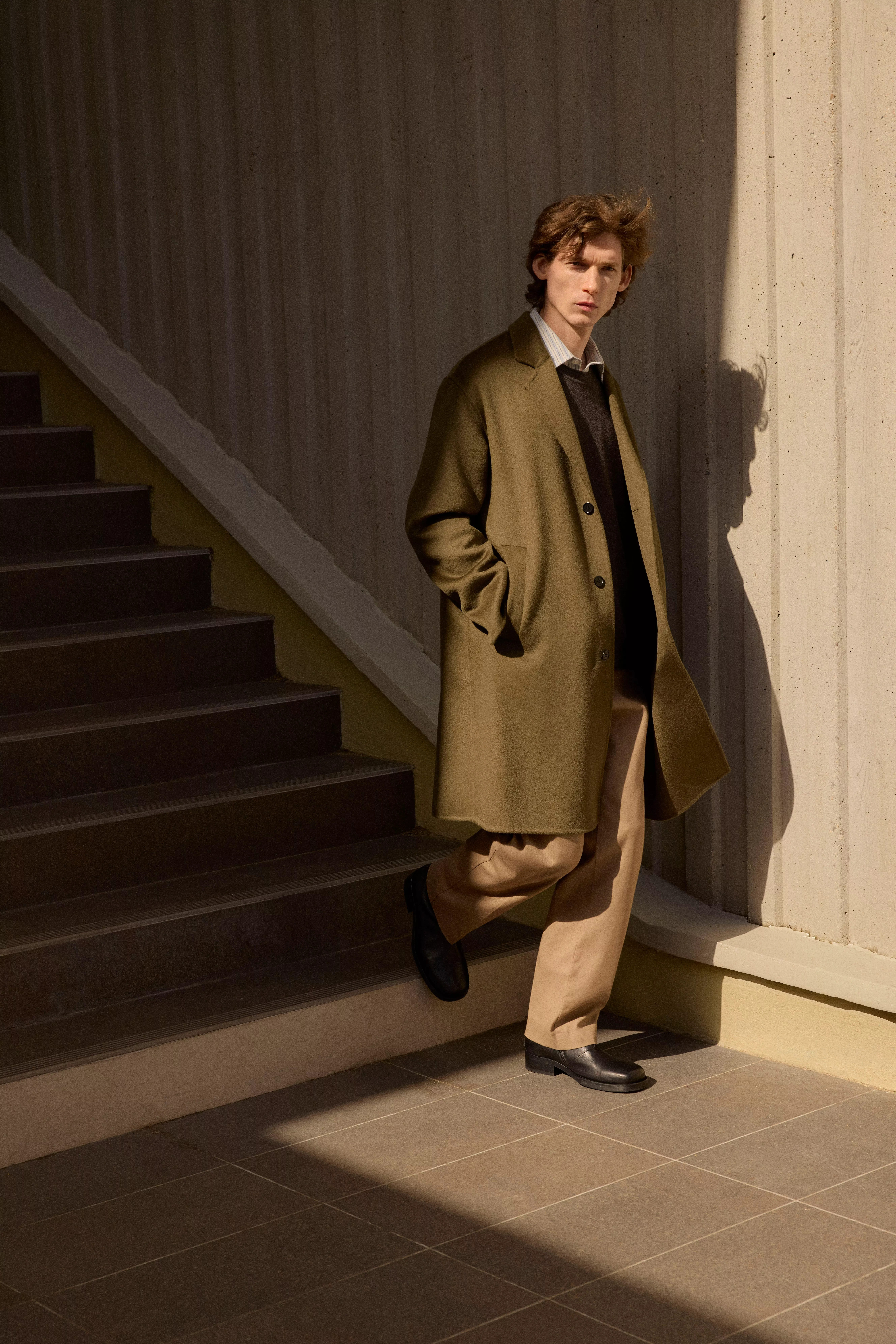 Menswear fashion model in olive overcoat and beige trousers by concrete stairs in warm sunlight outdoors