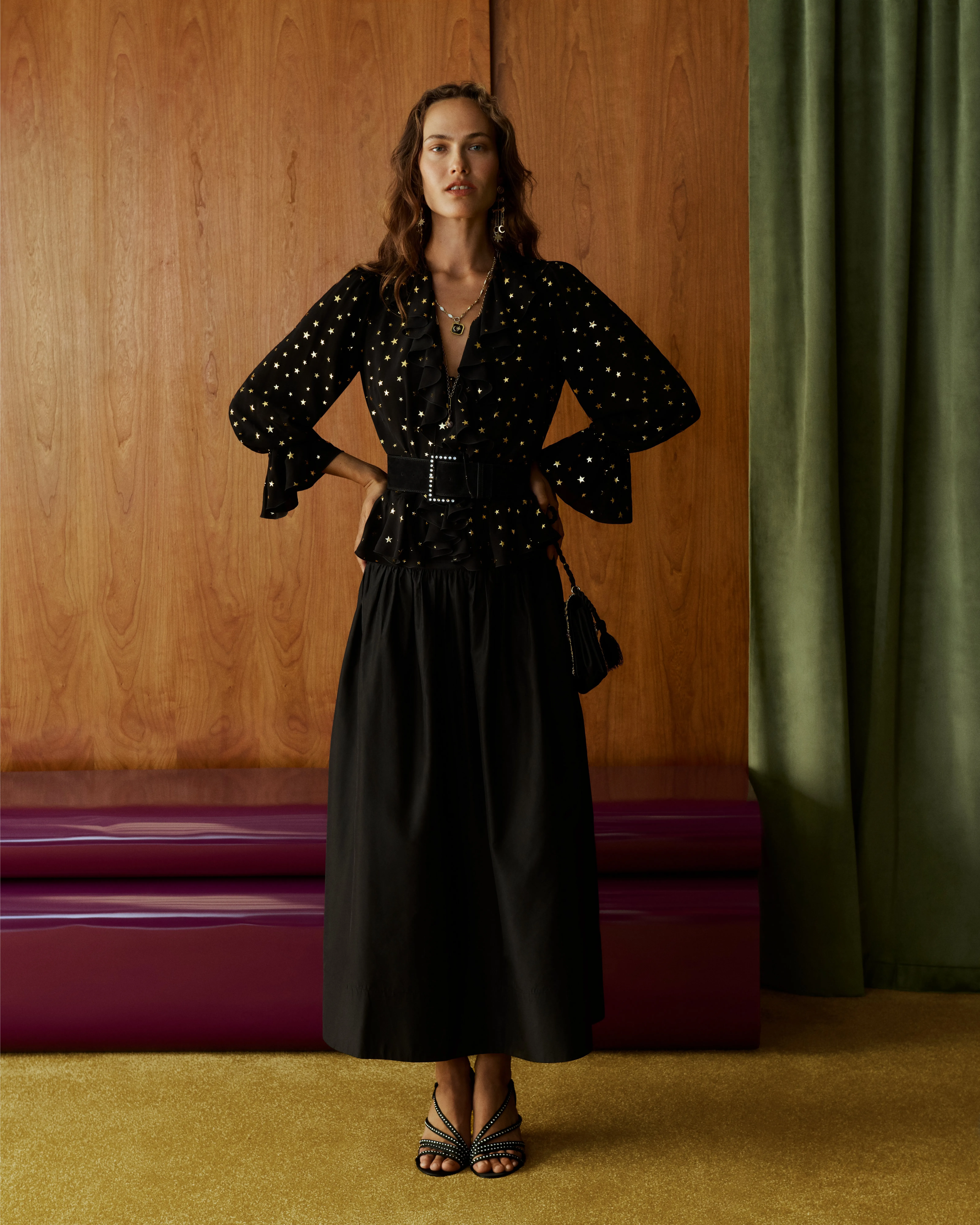 Model in black star-print blouse and maxi skirt with belt, posing by wood wall and green curtain indoors