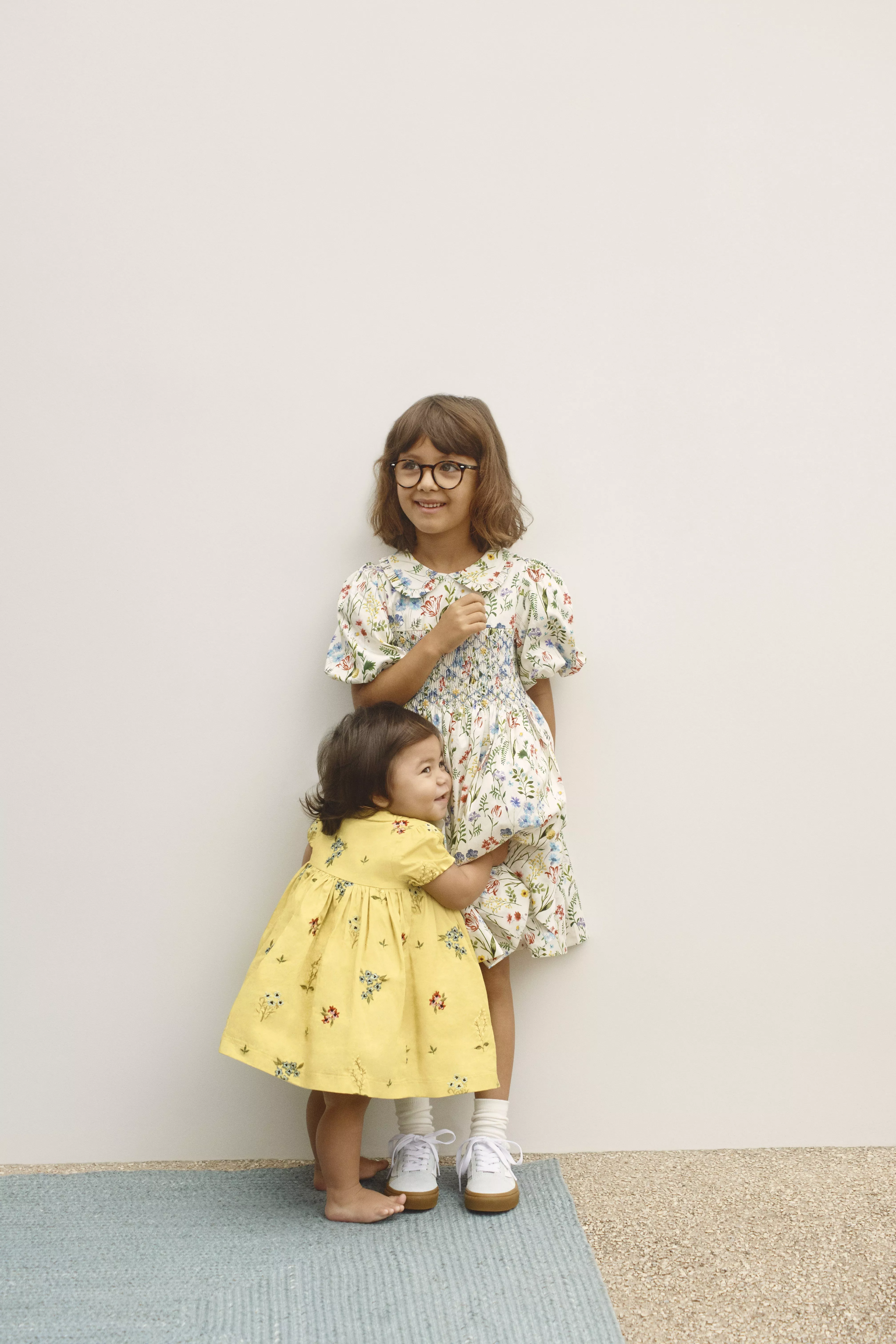 Toddler in a yellow dress hugging girl in a cream floral dress