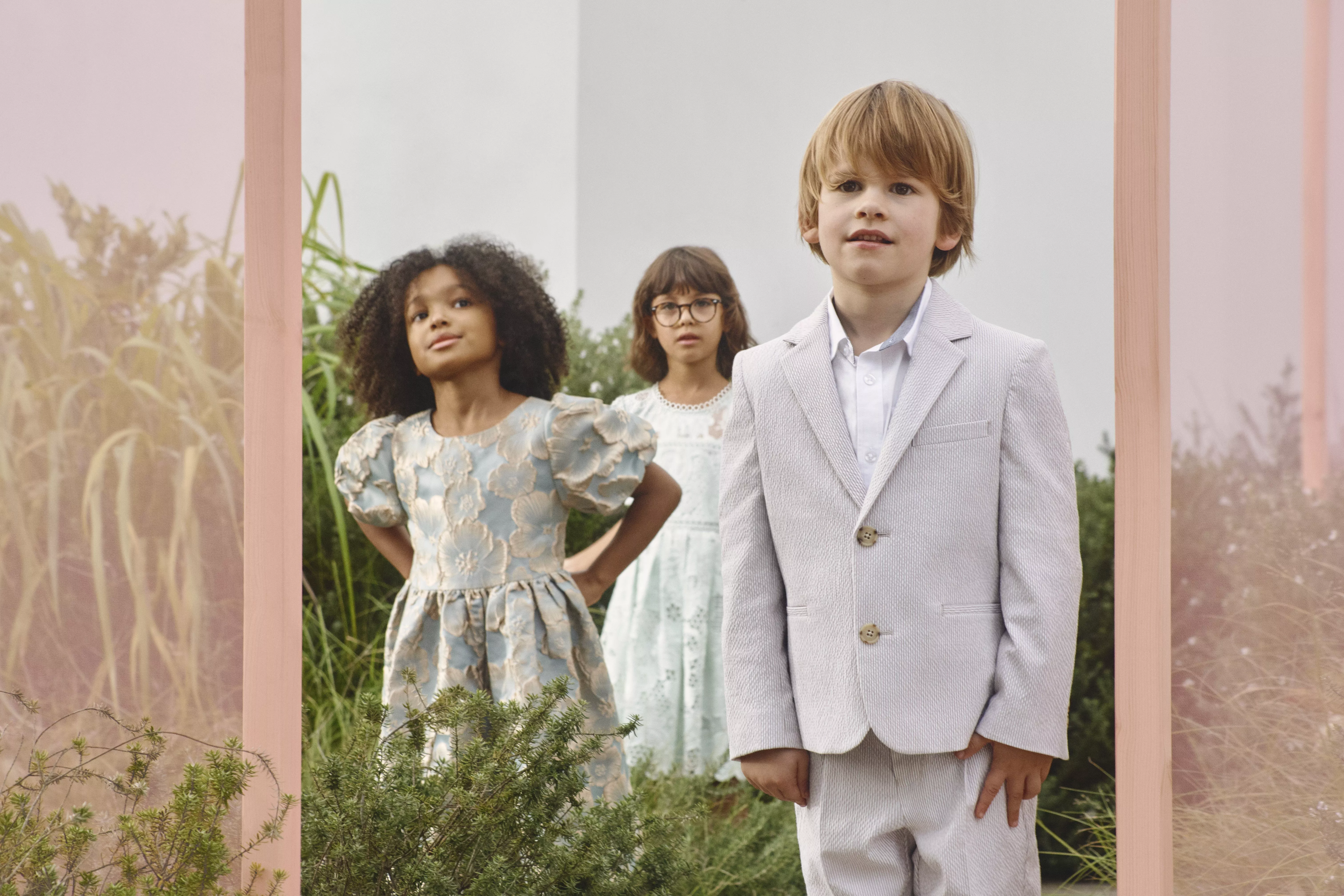 A boy wearing a light grey suit, with two girls wearing floral dresses standing behind him 