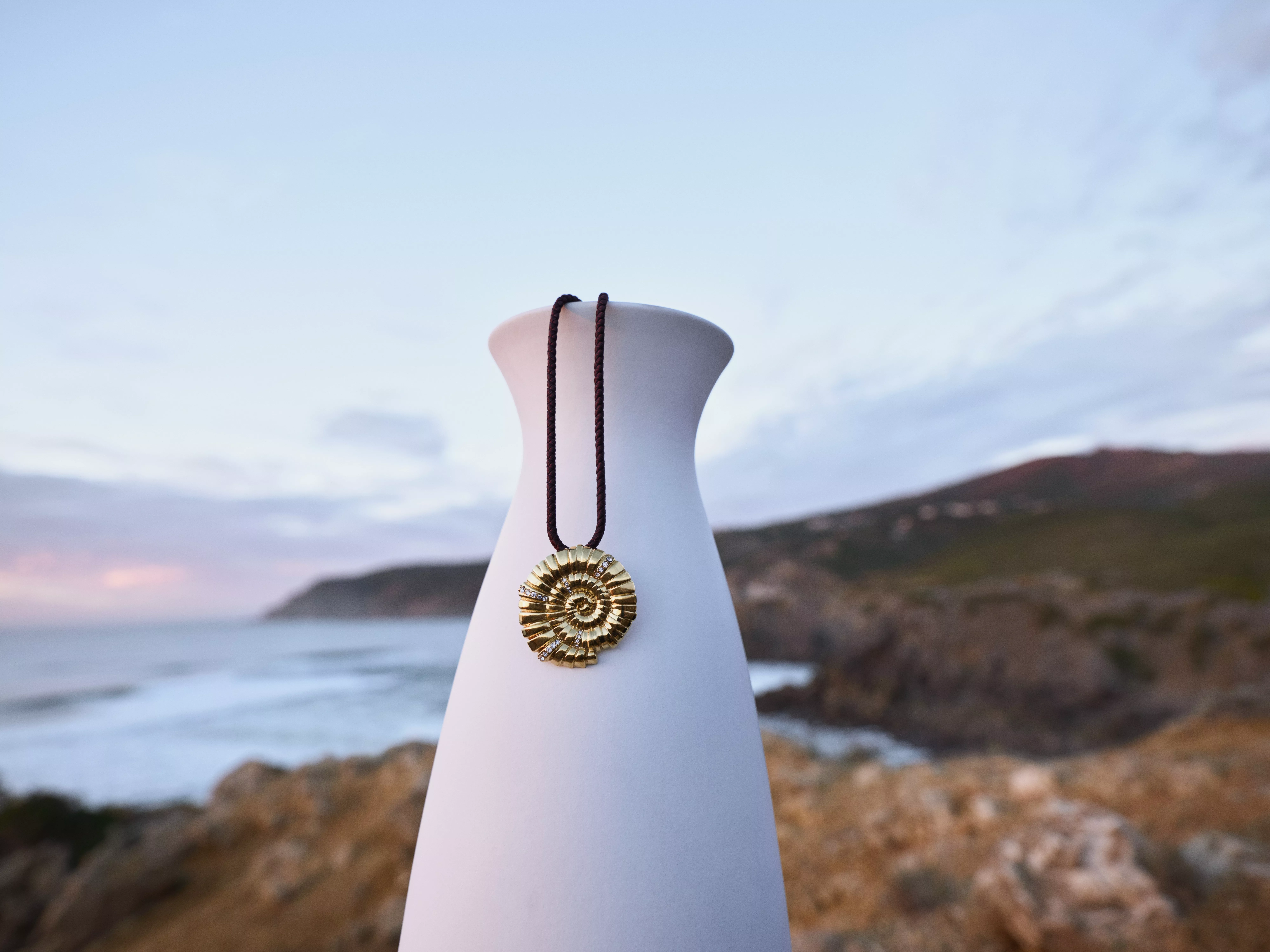 Gold spiral pendant necklace on brown cord, resting on a white vase