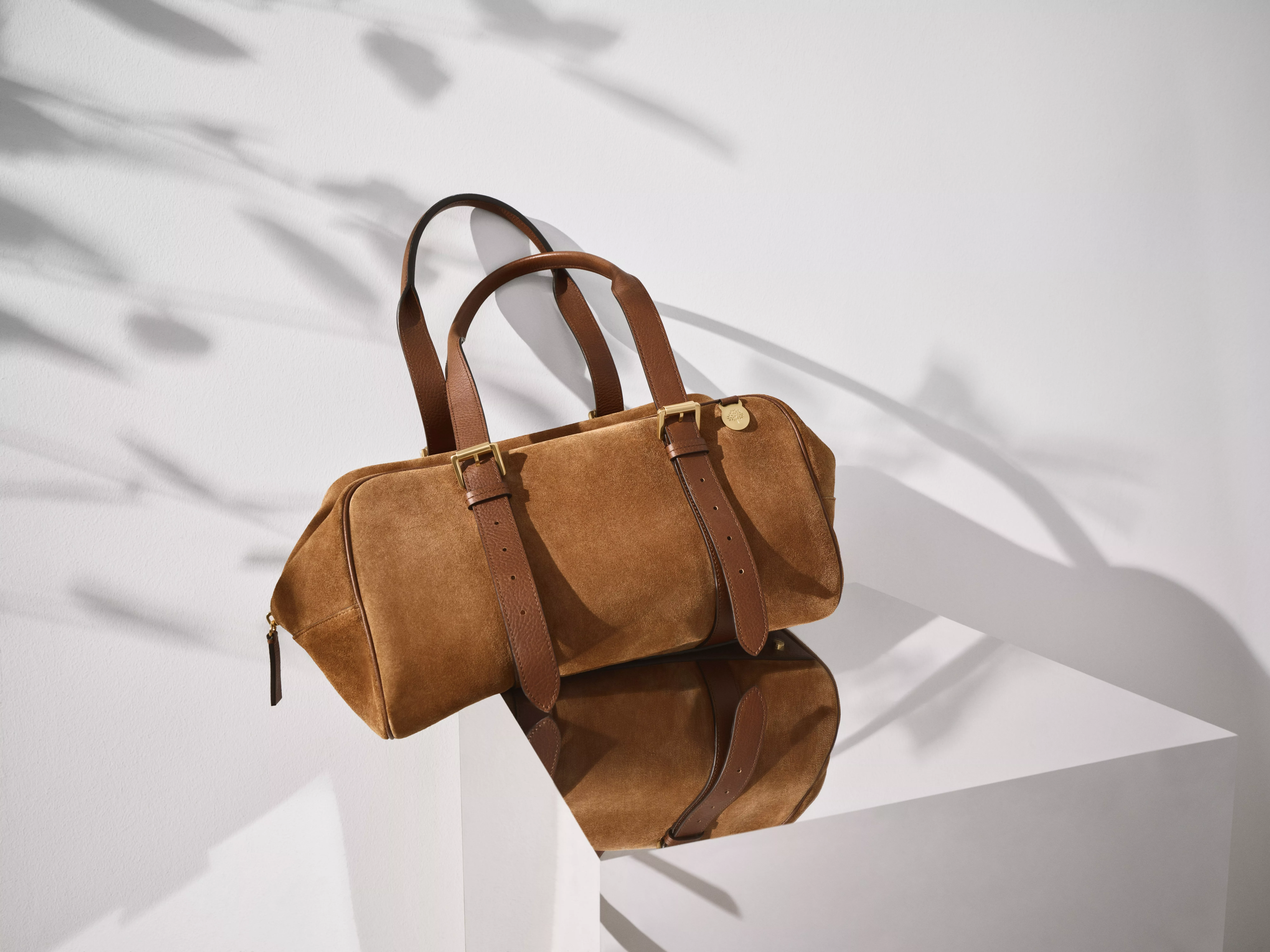 Elegant brown suede bag with leather straps and gold hardware, casting a shadow on a reflective surface.