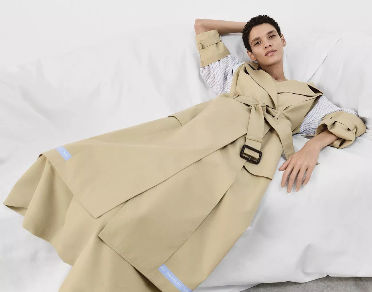 Person reclining on a white sofa, wearing a stylish beige trench coat with a belt and rolled-up sleeves.