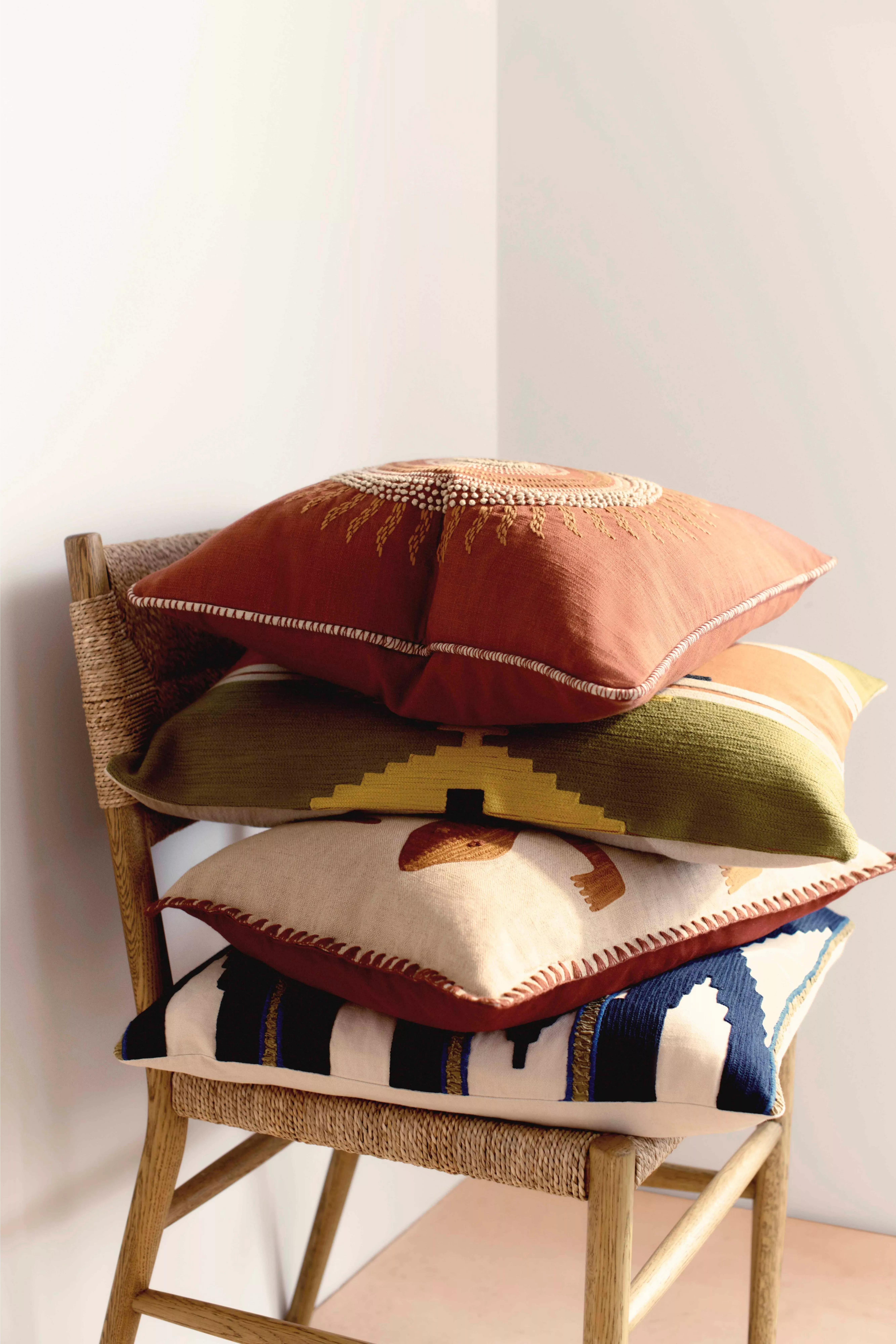 A stack of four embroidered cushions with geometric patterns in earthy tones, resting on a woven chair.