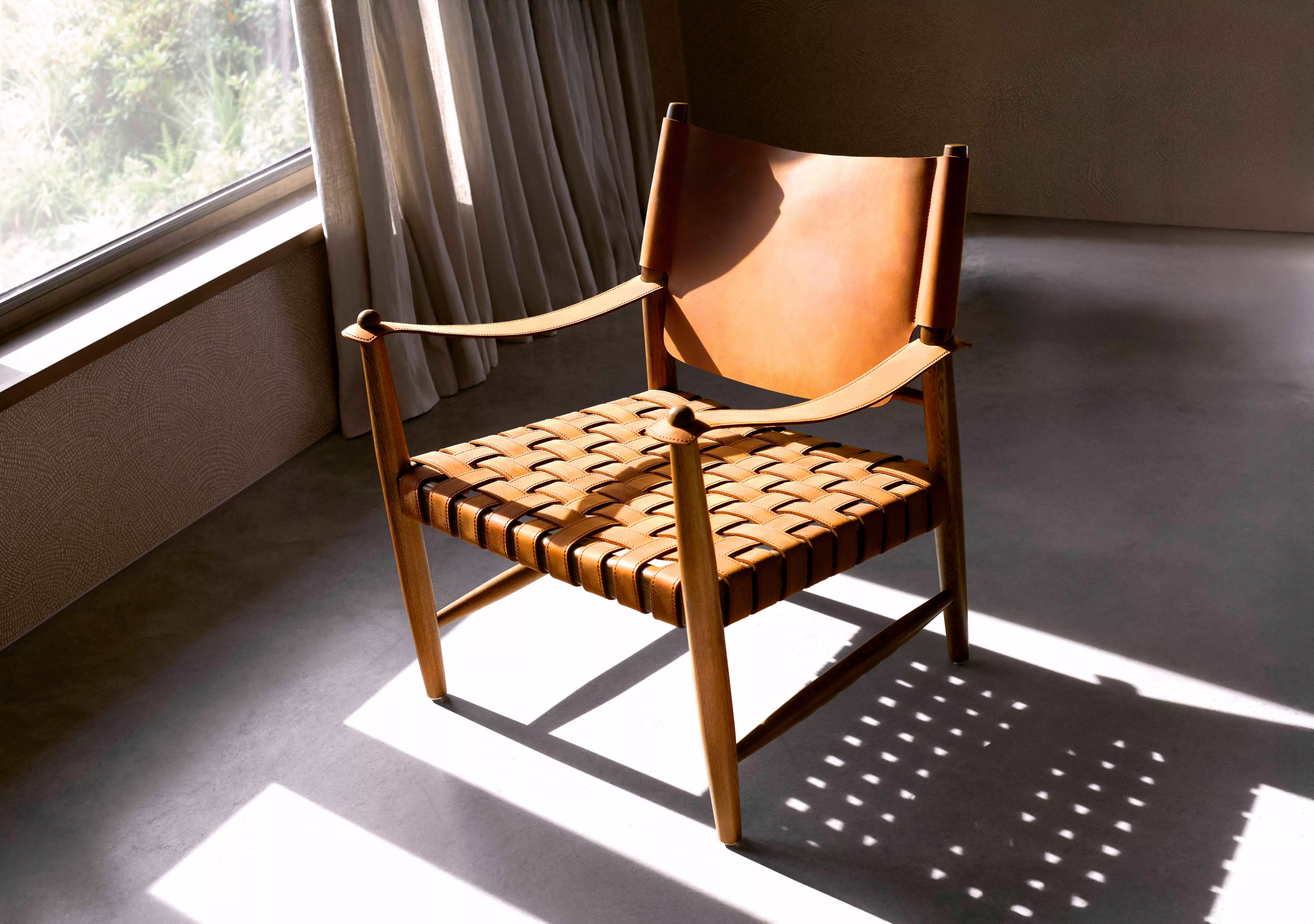Stylish wooden chair with woven leather seat and backrest, bathed in sunlight near a window with curtains.