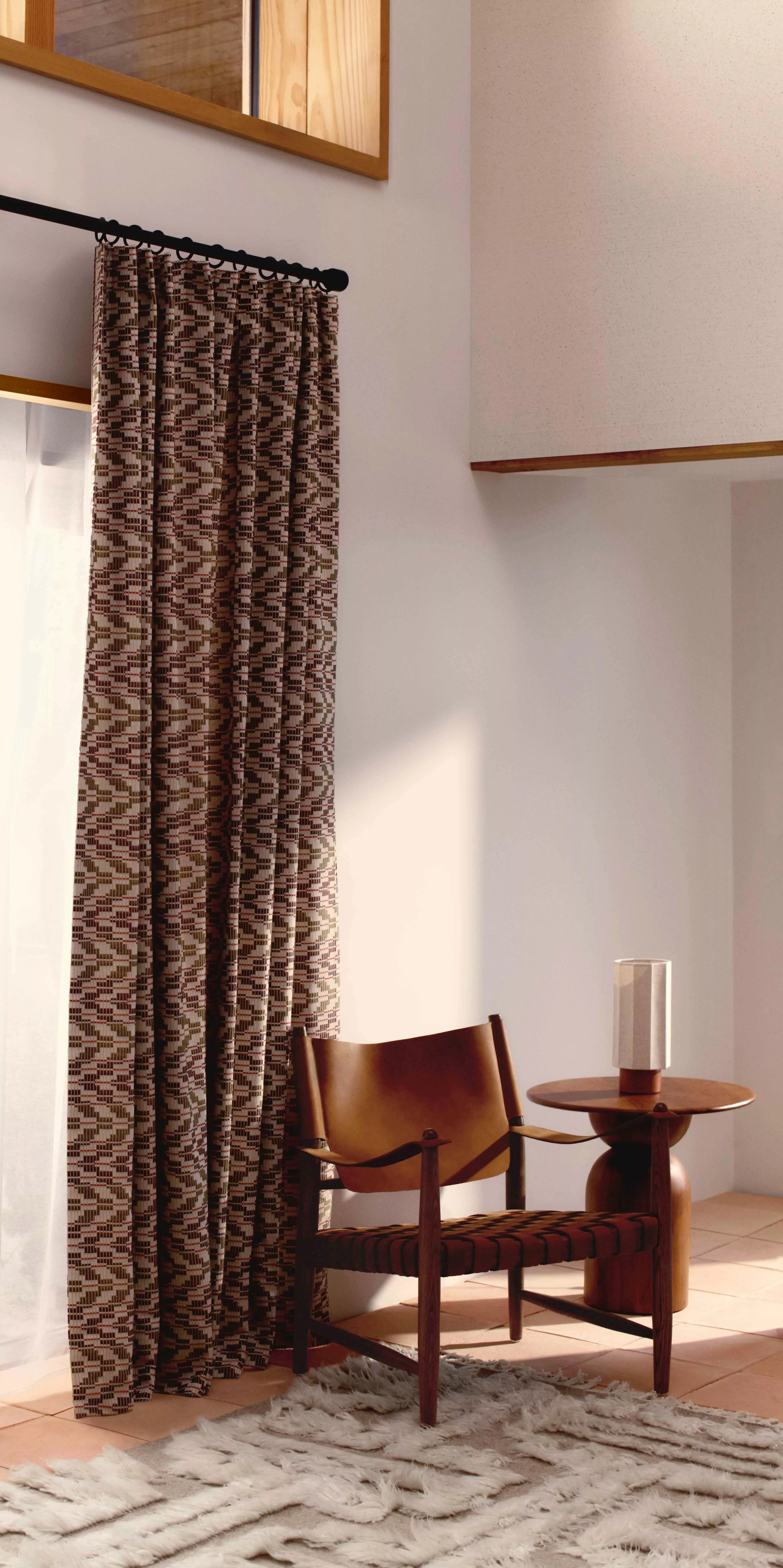 Cosy room with patterned curtains, a wooden chair, a round table, and a textured rug on tiled floor.