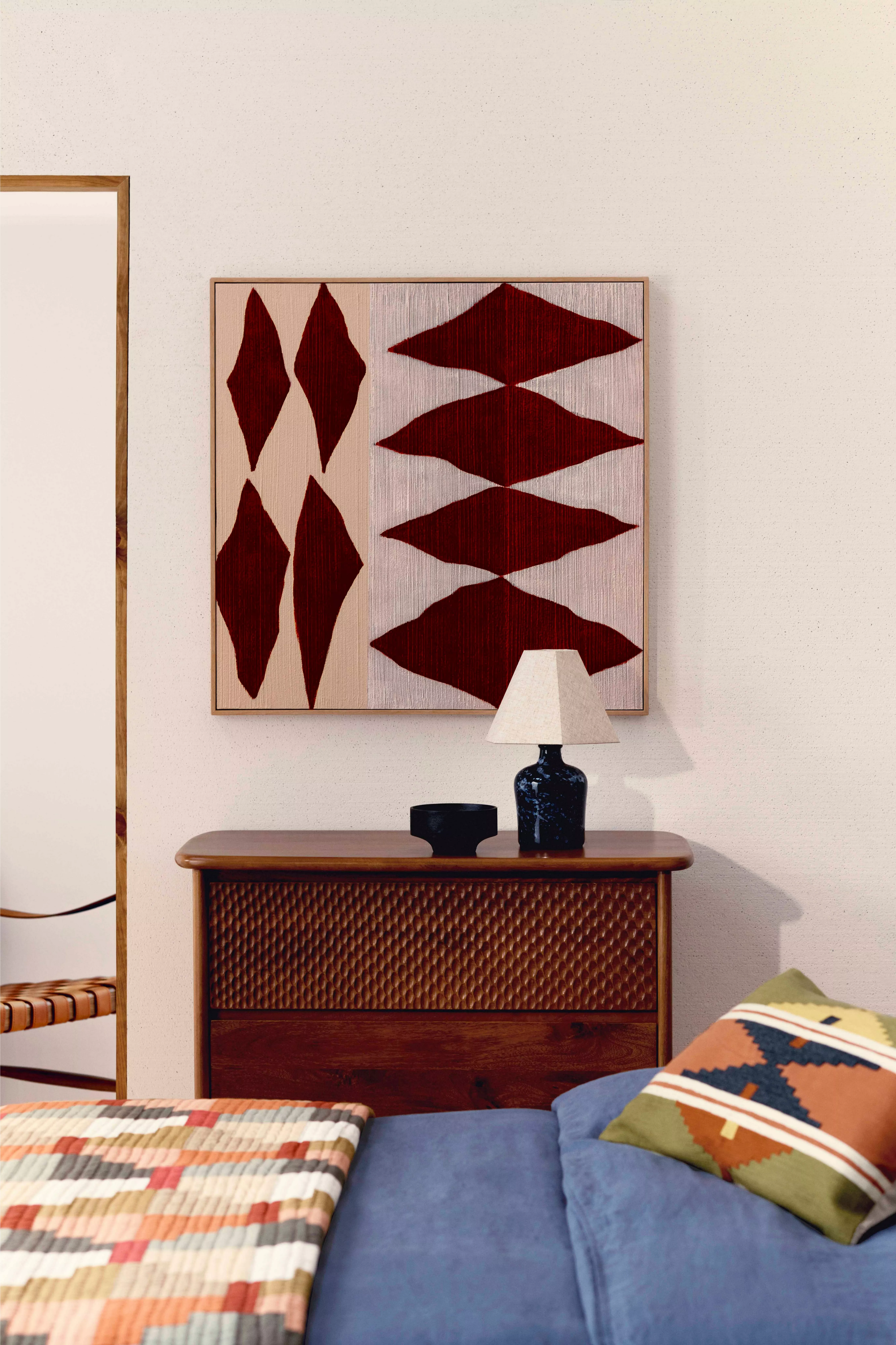Modern bedroom with geometric wall art, wooden dresser, and textured quilt. Stylish lamp and cushion accents.