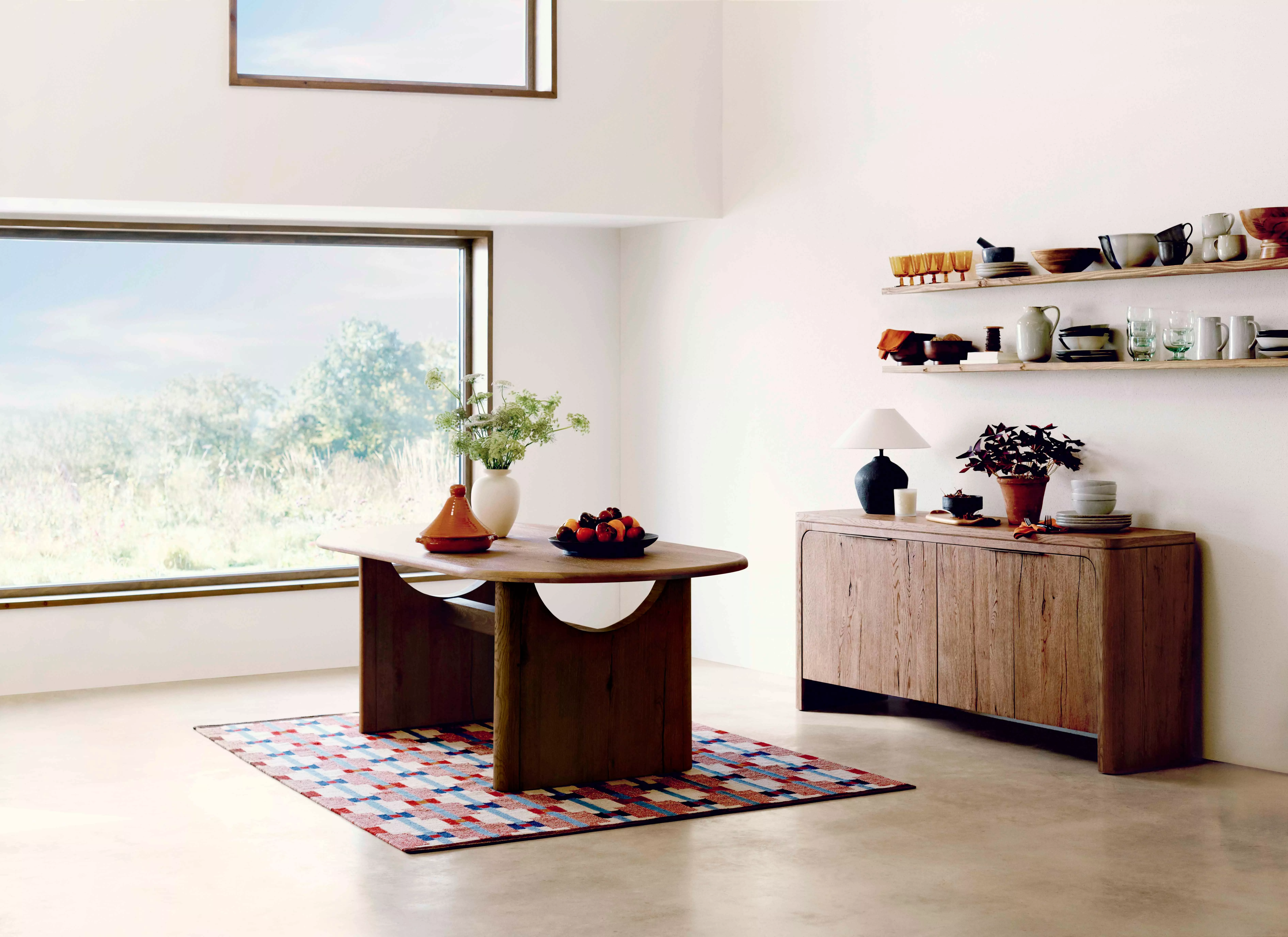 Modern dining room with a wooden table, colourful rug, and large windows. Minimalist decor on shelves.