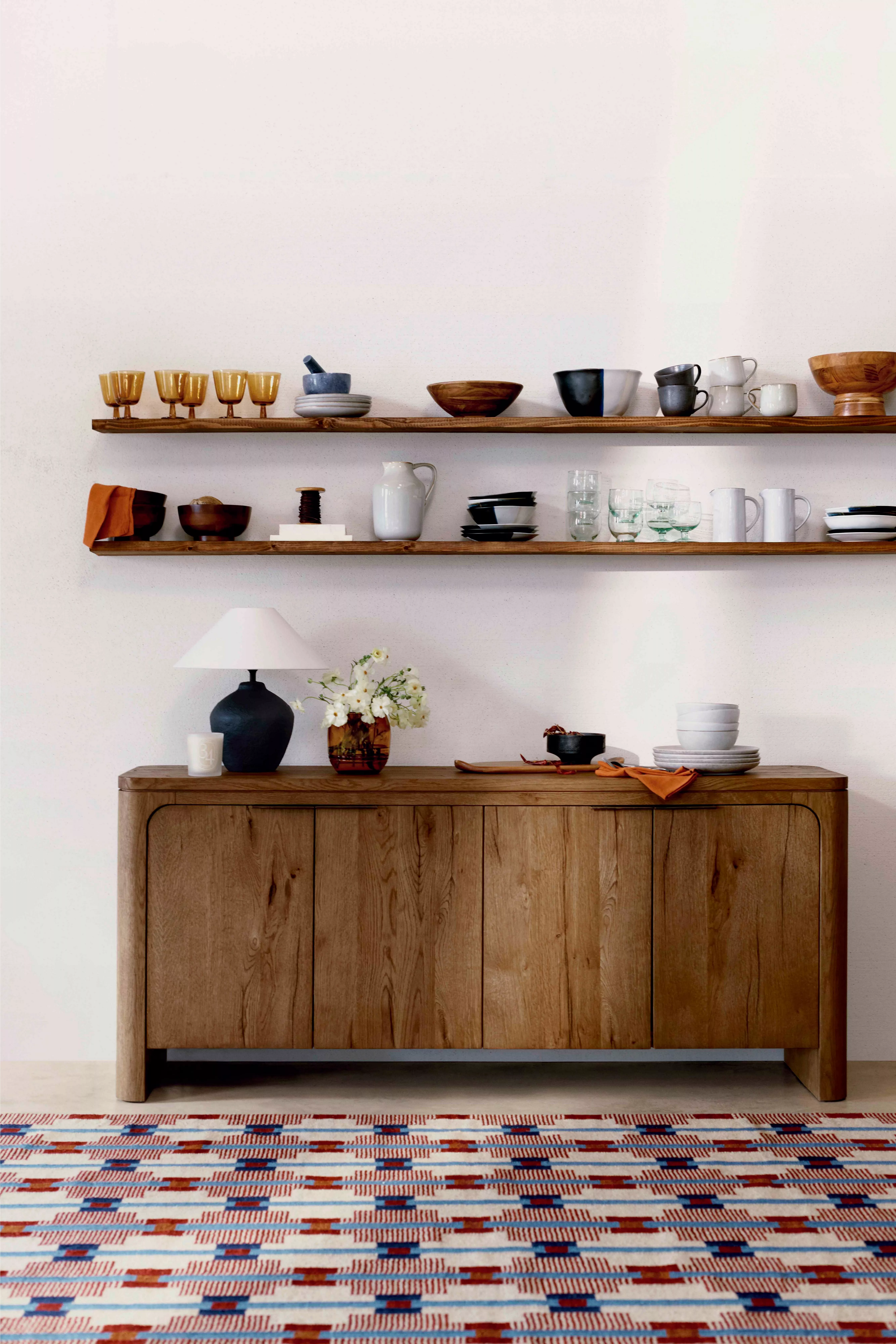 A light oak console table with three drawers and a woven shelf, styled with a tall vase of greenery, a lamp, and books.