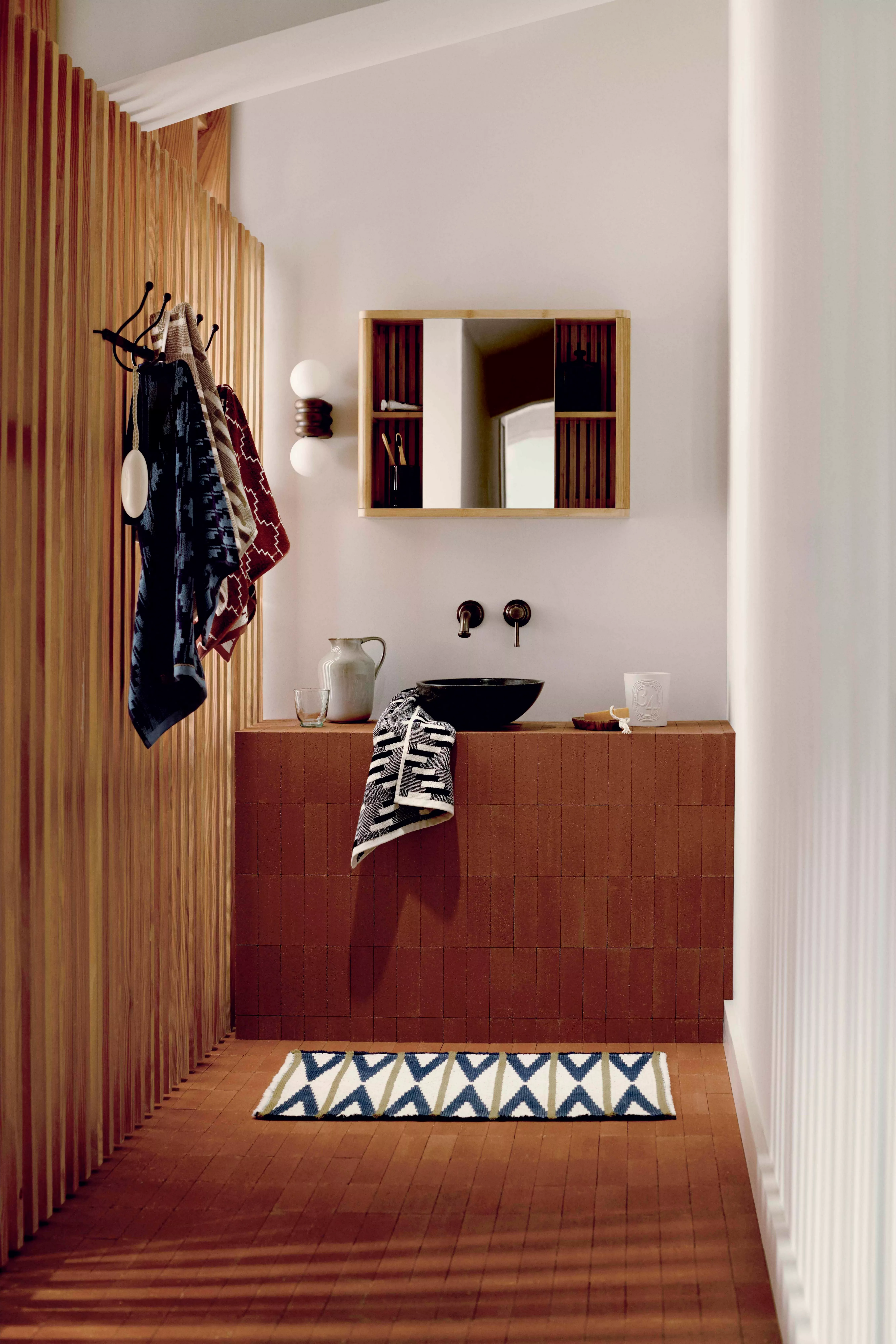 Modern bathroom with terracotta tiles, wooden accents, and geometric textiles. Wall-mounted sink and mirror.