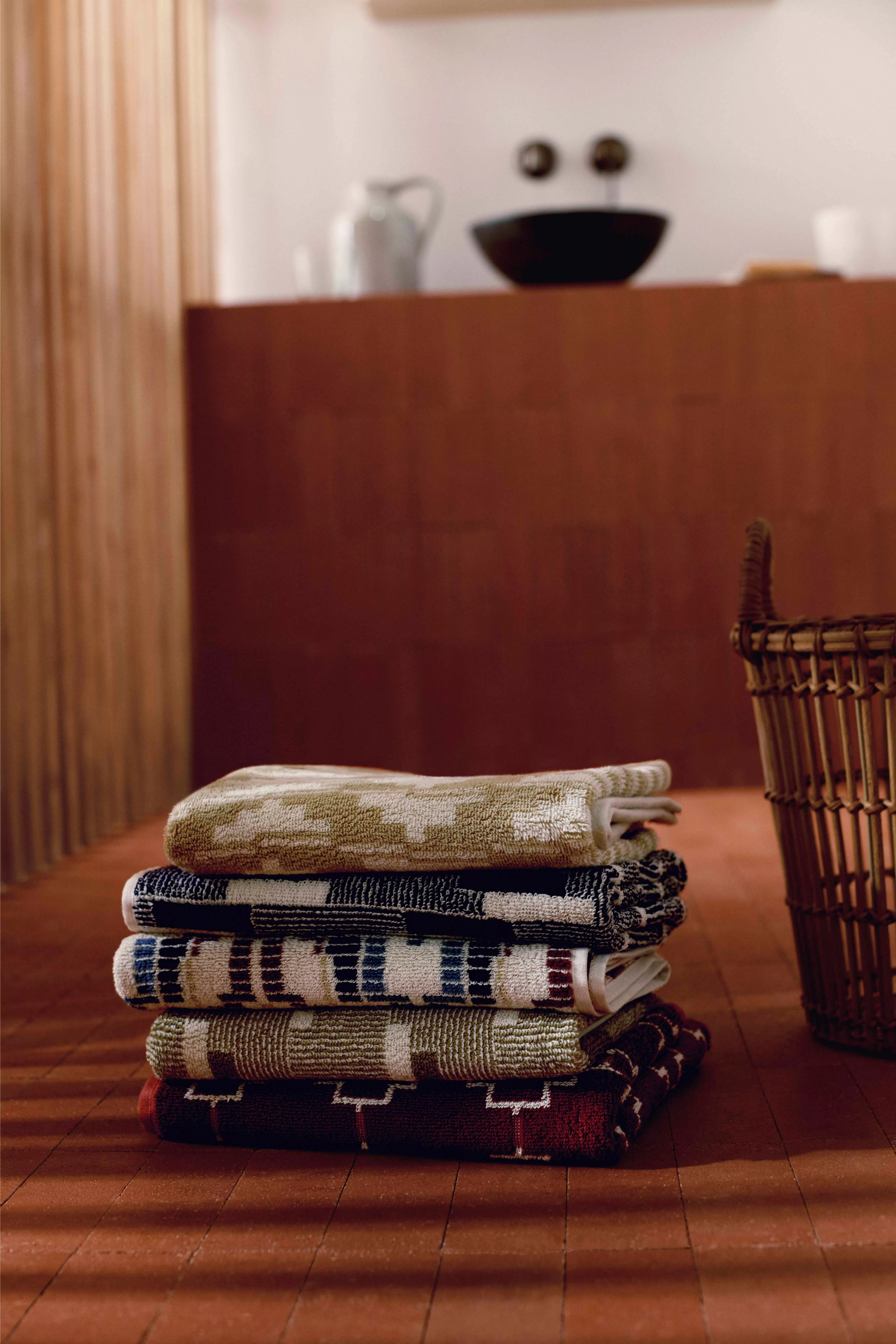Stack of folded patterned towels on a terracotta floor, with a wicker basket nearby in a cosy bathroom setting.