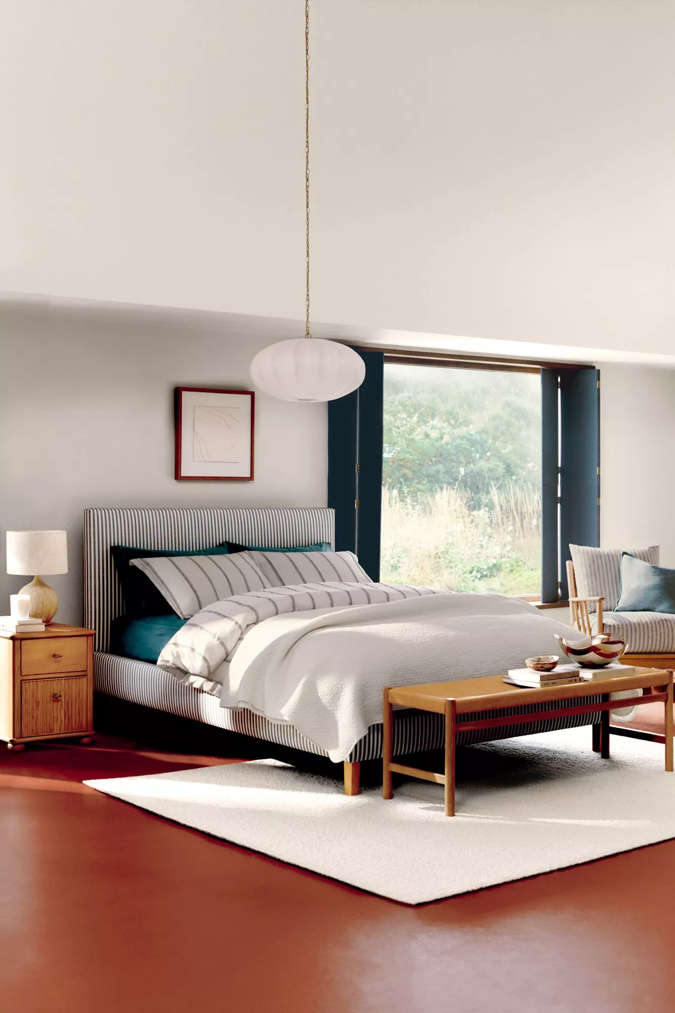 Minimalist bedroom with striped bedding, wooden furniture, and a large window with a garden view.