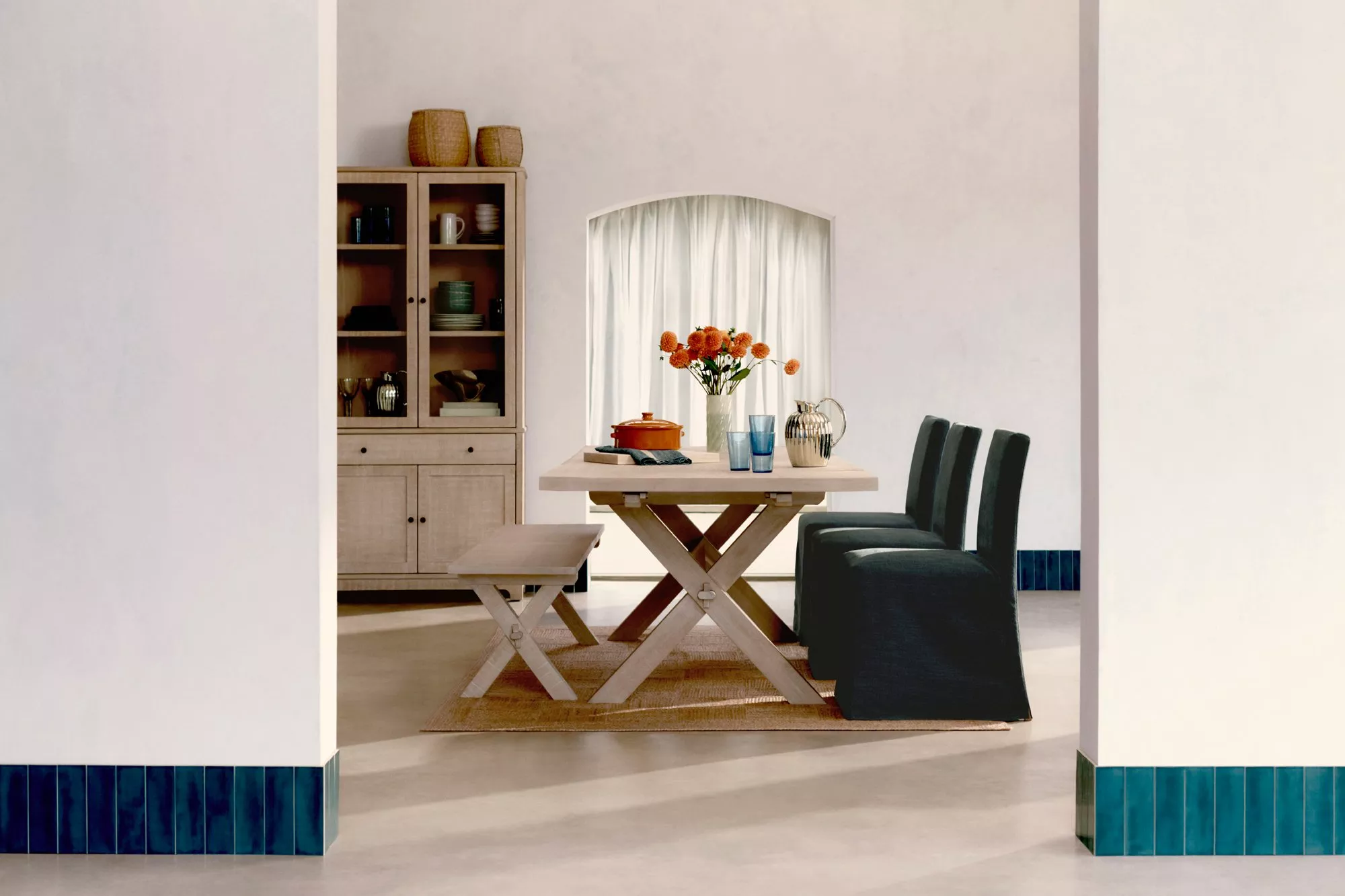 Modern dining room with a wooden table, orange flowers, and dark chairs. Shelves with dishes in the background.