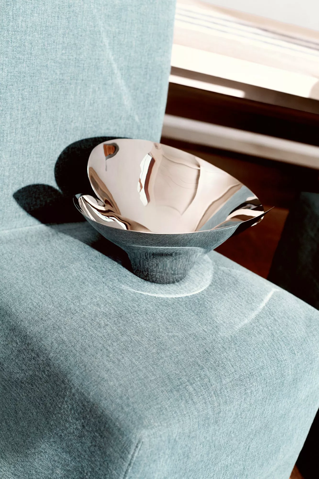 A reflective silver bowl sits on a light blue upholstered chair, casting shadows and reflecting its surroundings.
