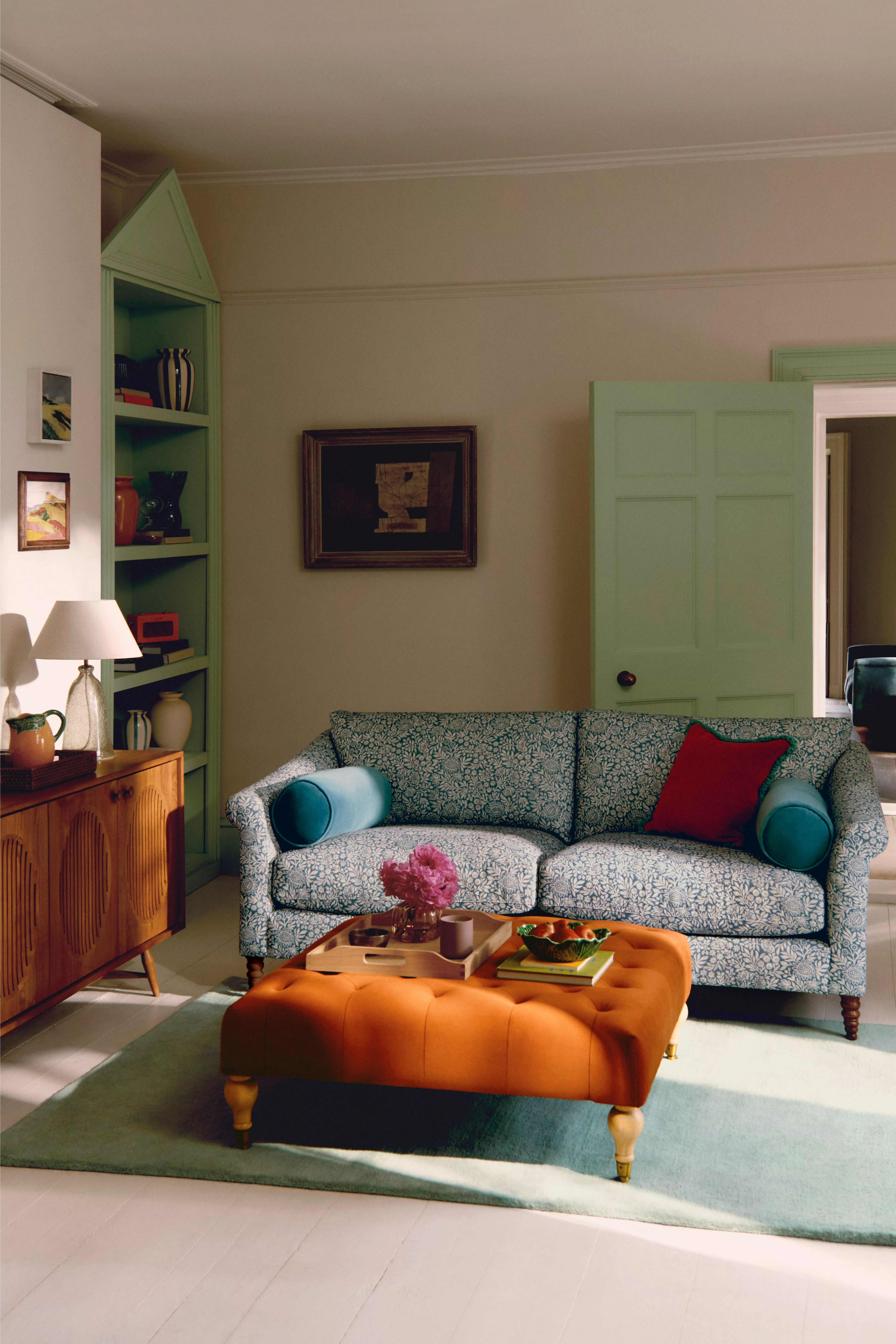 Stylish living room with patterned sofa, orange tufted ottoman, green shelving and pastel door decor