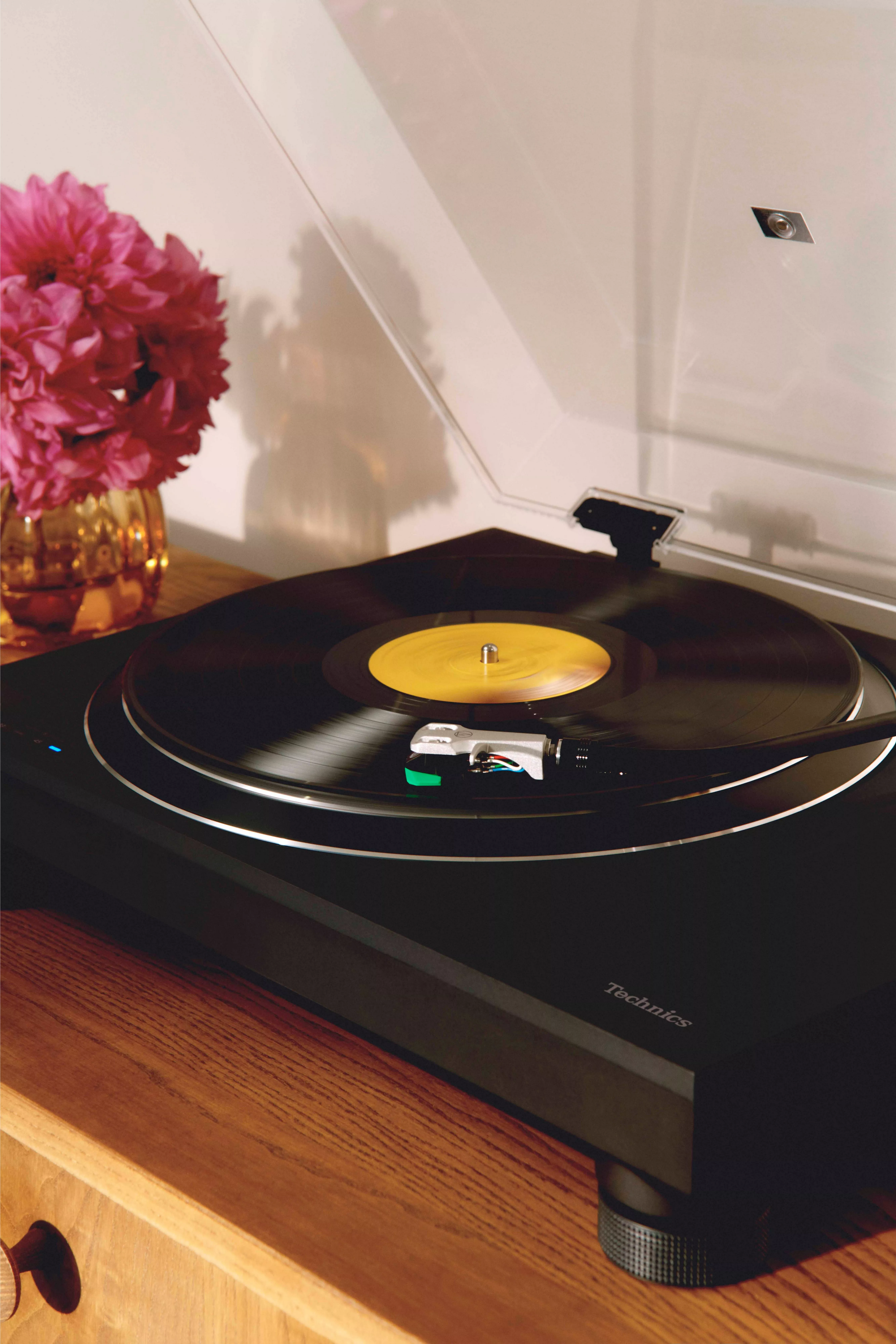 Technics turntable playing a vinyl record on a wooden cabinet, with pink flowers in a vase beside it