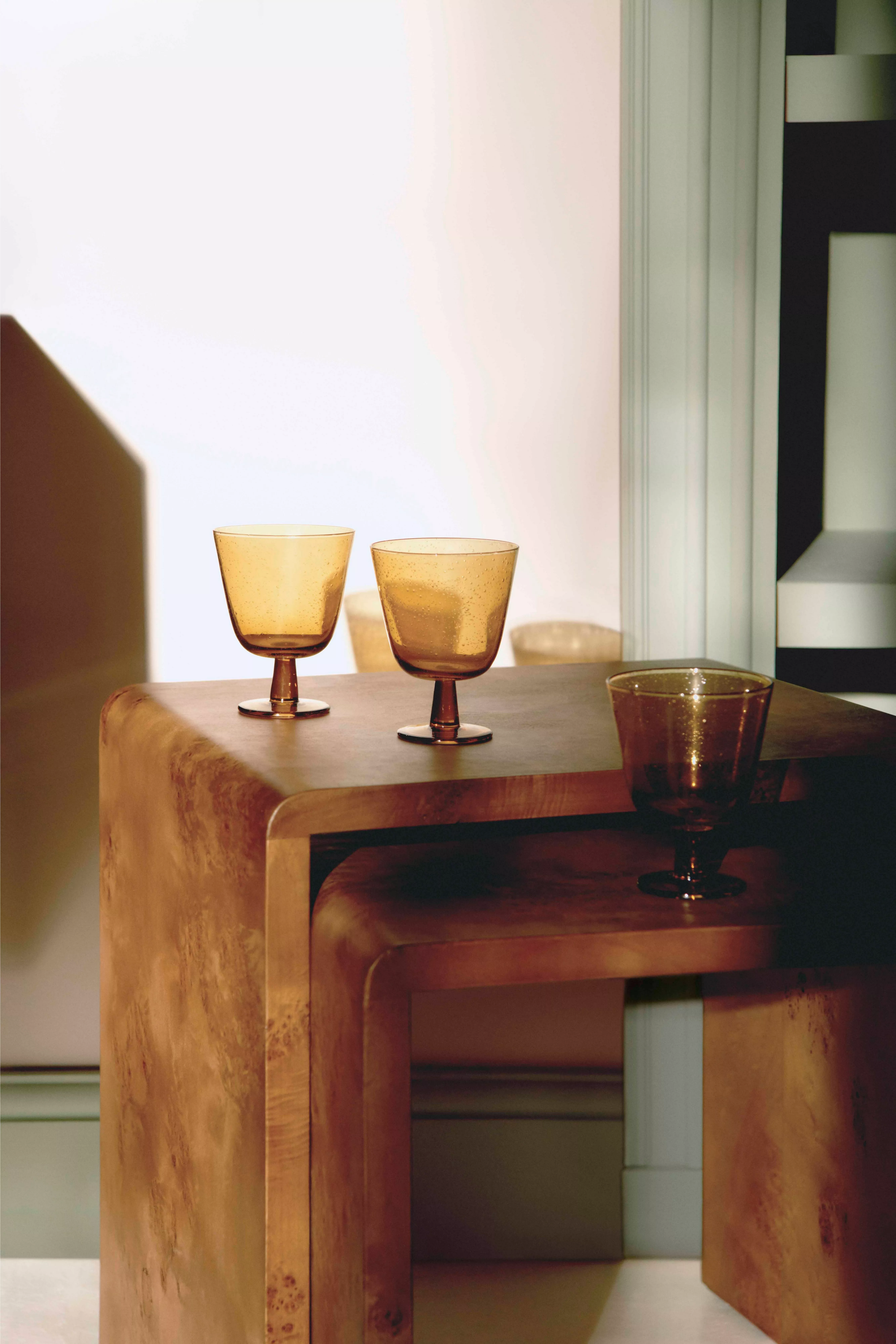 Amber glass goblets on sculptural wooden side tables in warm sunlight, minimalist modern interior décor scene