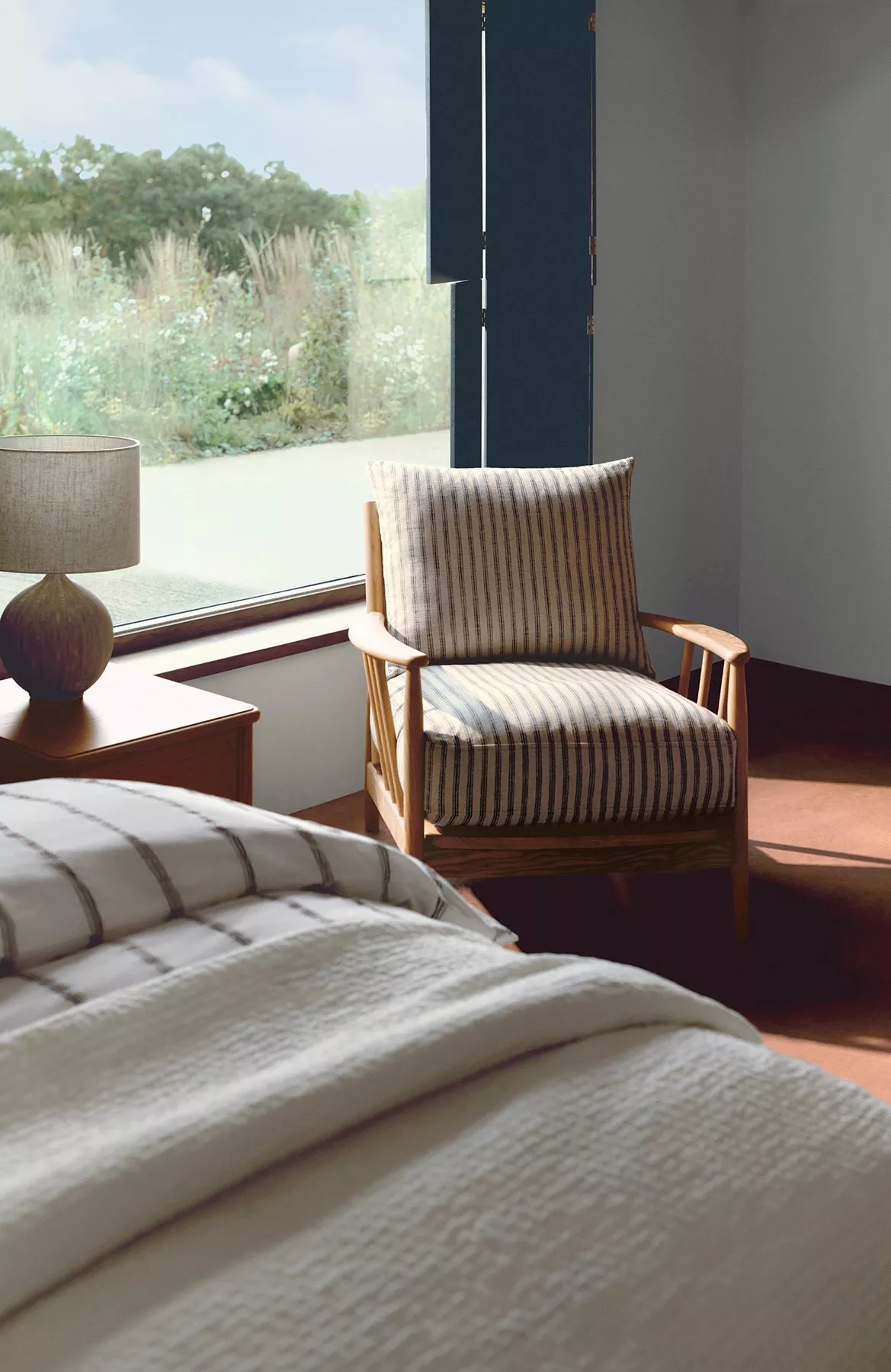 Cosy bedroom with a striped armchair by a large window, neutral tones, and a soft bedspread in natural light.