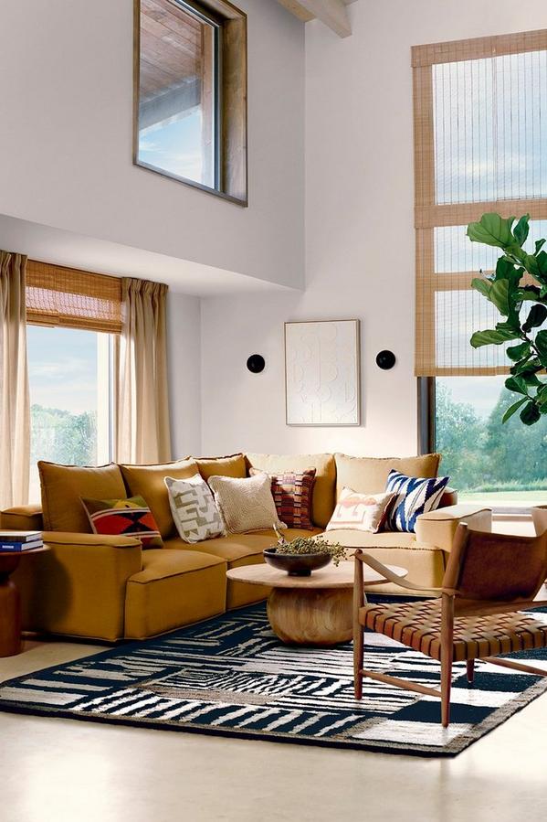 Modern living room with mustard sofa, patterned cushions, wooden table, and large windows overlooking greenery.