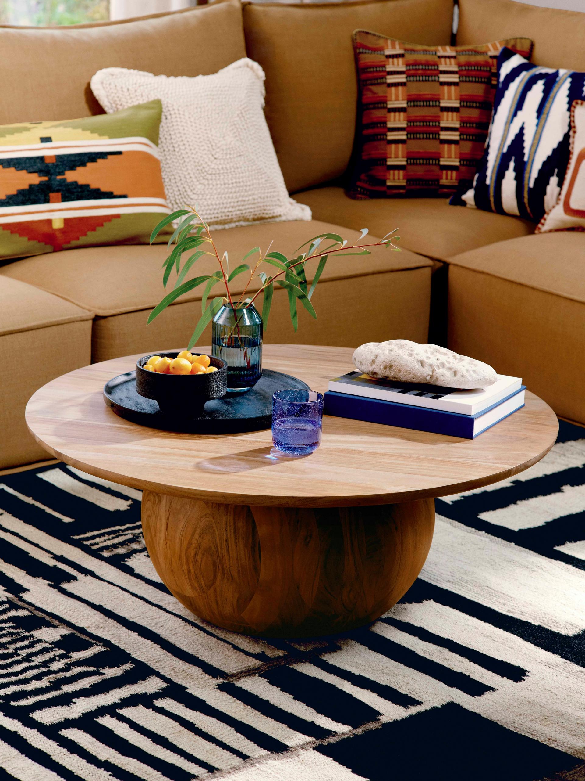 Round wooden coffee table on black and white rug, styled with tray, vase, books, beside tan sectional sofa