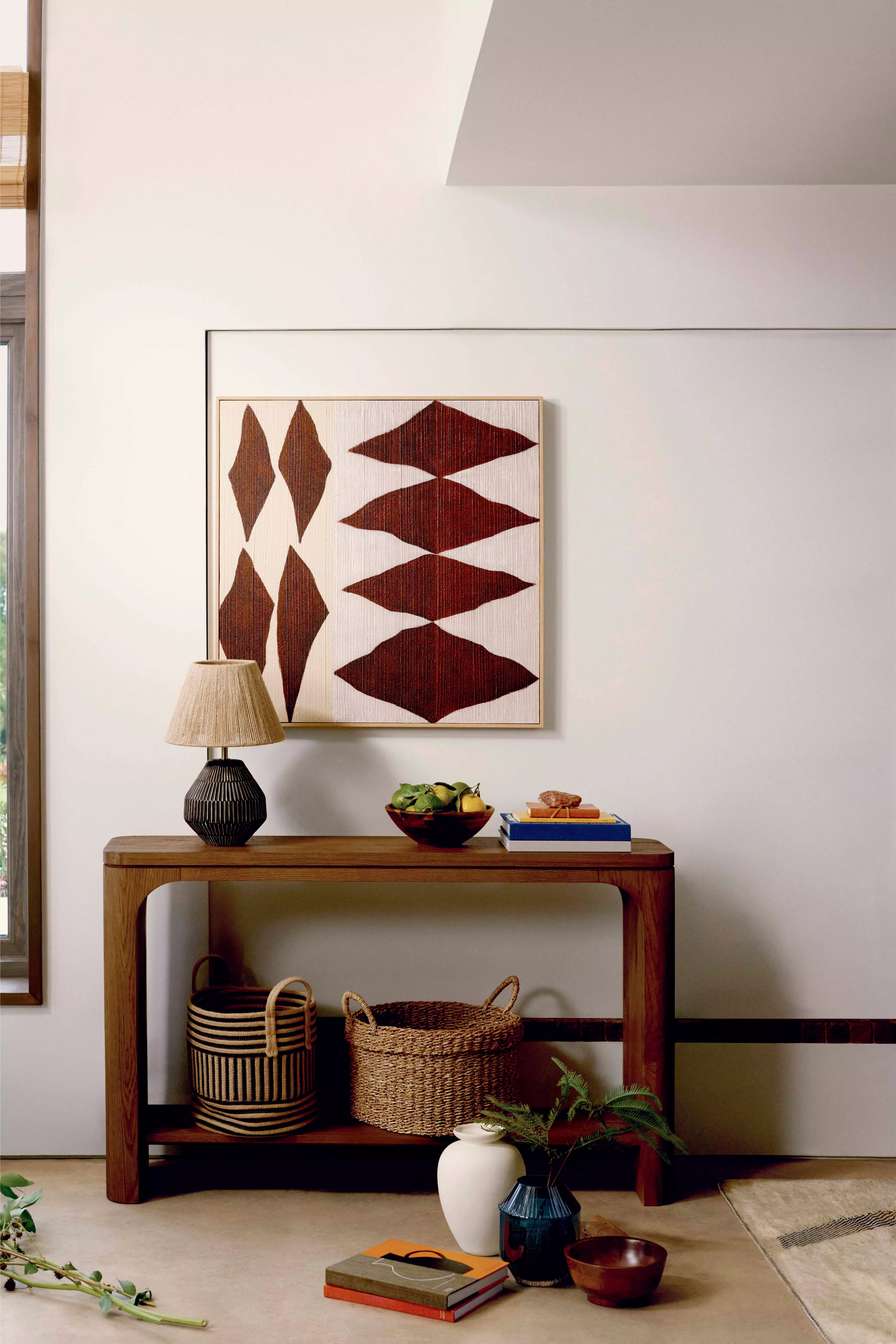 A light oak console table with three drawers and a woven shelf, styled with a tall vase of greenery, a lamp, and books.
