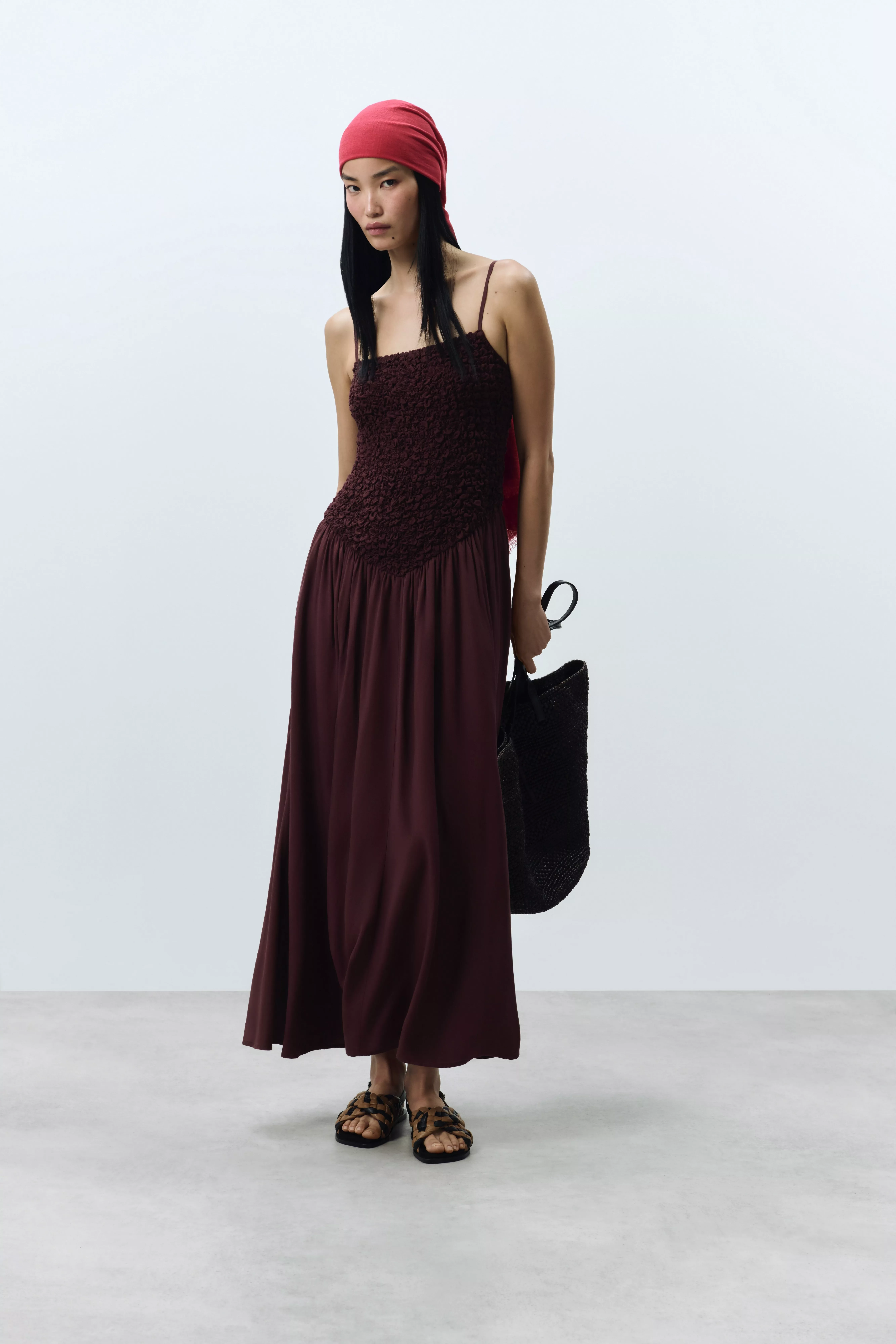 Woman in burgundy maxi dress with red headscarf, sandals and black tote bag, minimalist studio backdrop