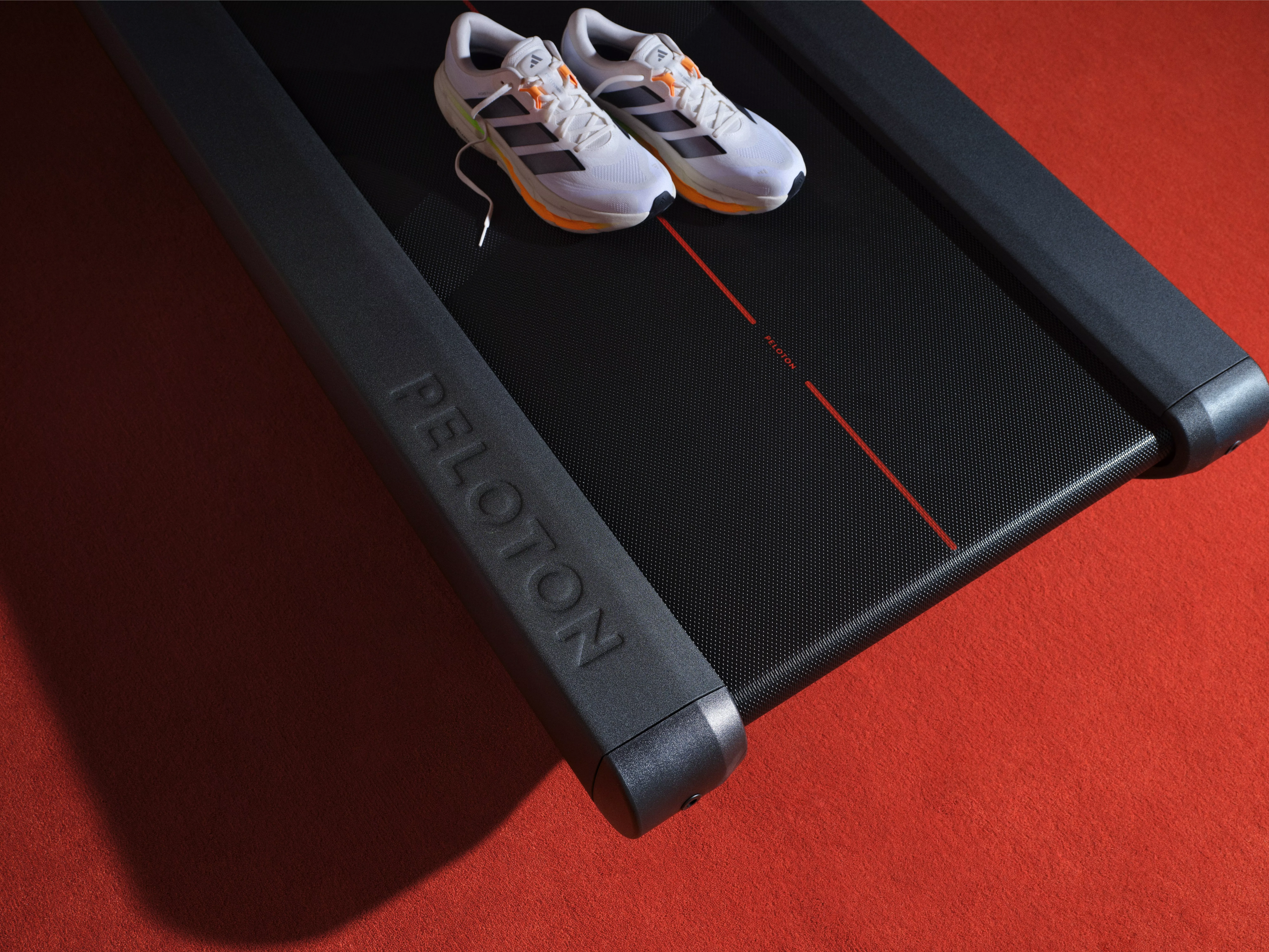 White running trainers on a Peloton treadmill belt in a gym, fitness equipment close-up on red floor.