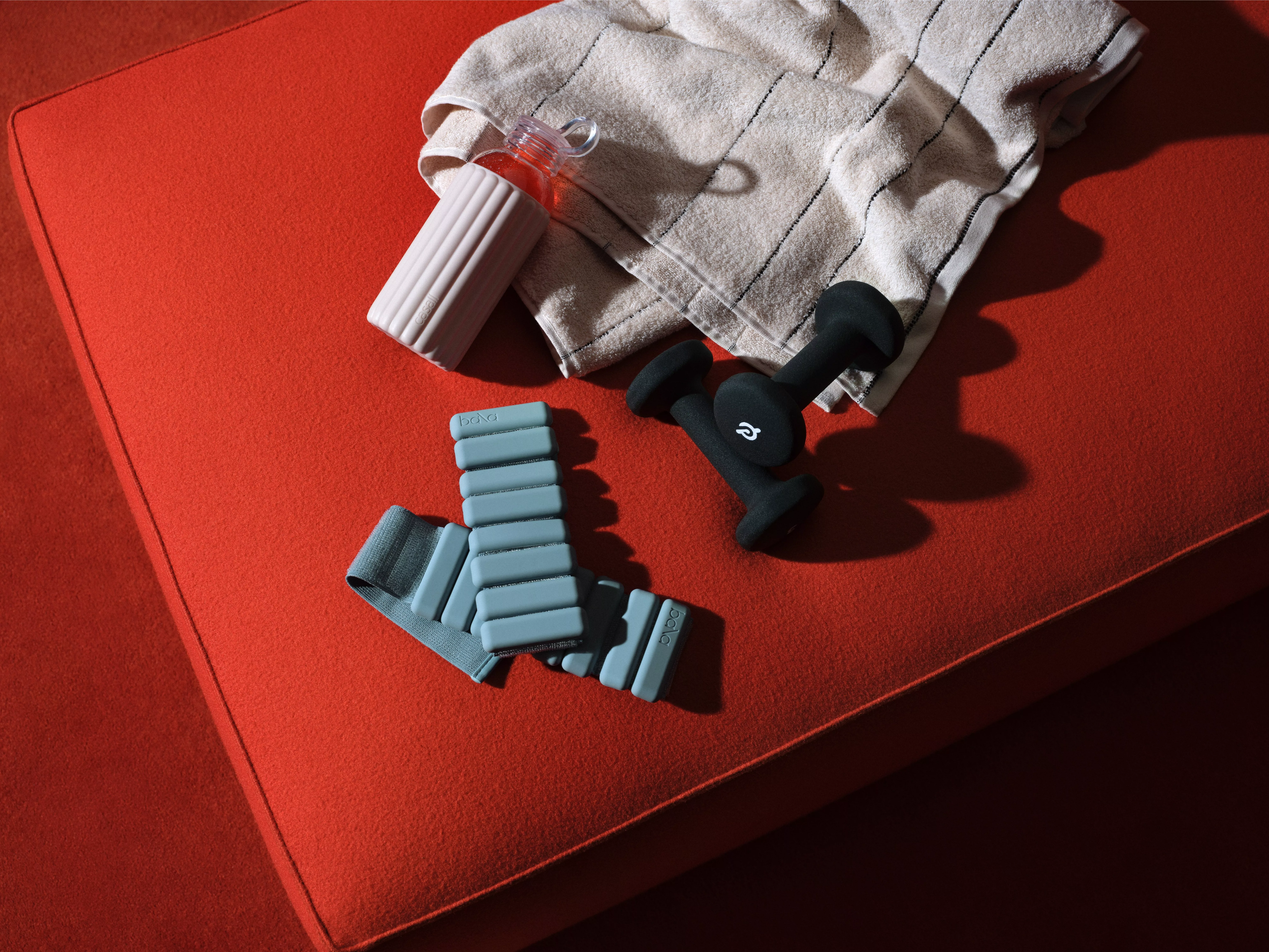 Fitness gear on a red yoga mat: dumbbells, ankle weights, water bottle and striped towel, flat lay workout setup