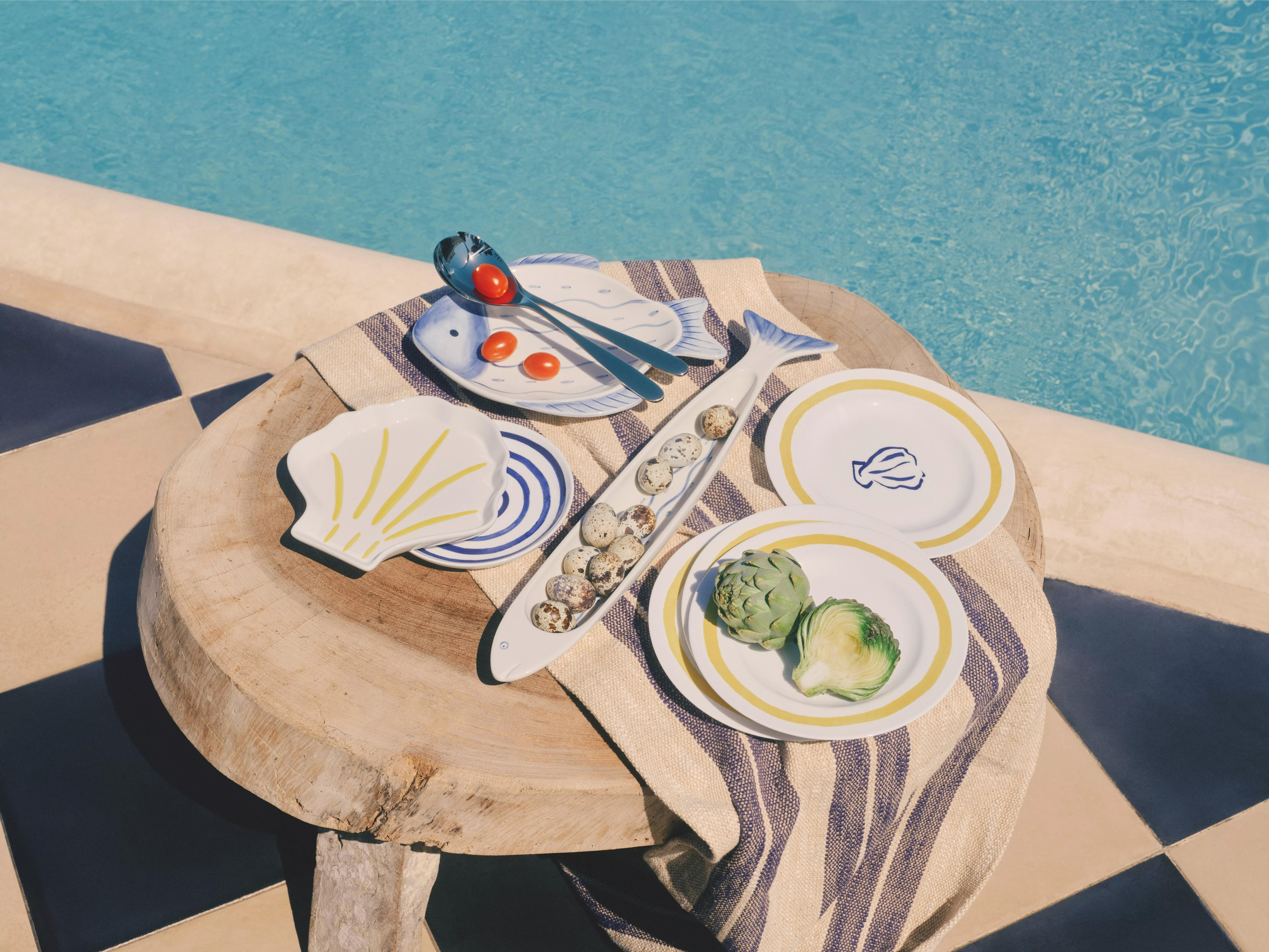 Poolside outdoor table setting with nautical ceramic plates, quail eggs, cherry tomatoes and artichoke on linen cloth